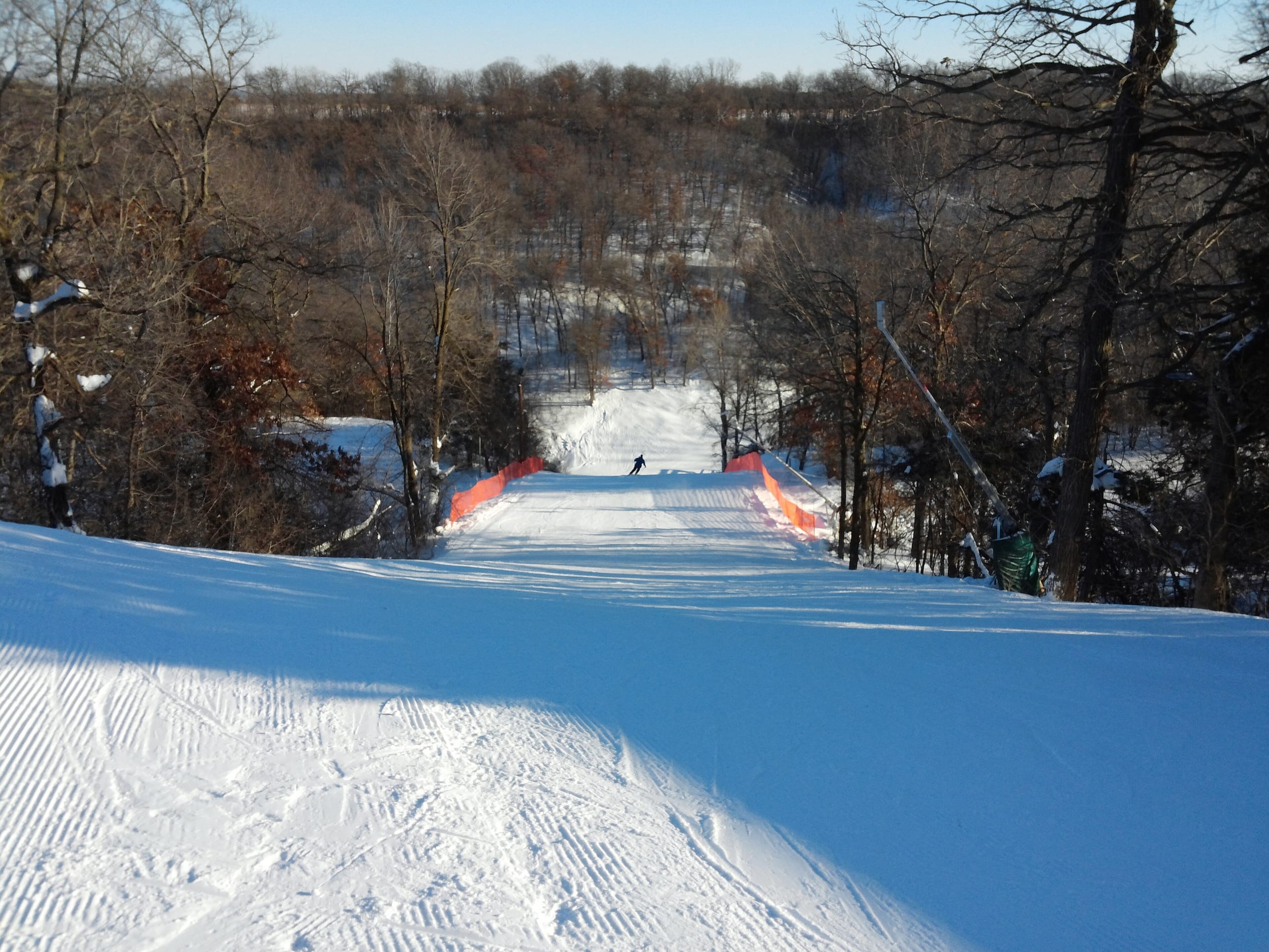 Groomed ski run lined with orange fencing dropping through bare winter trees, a lone skier below