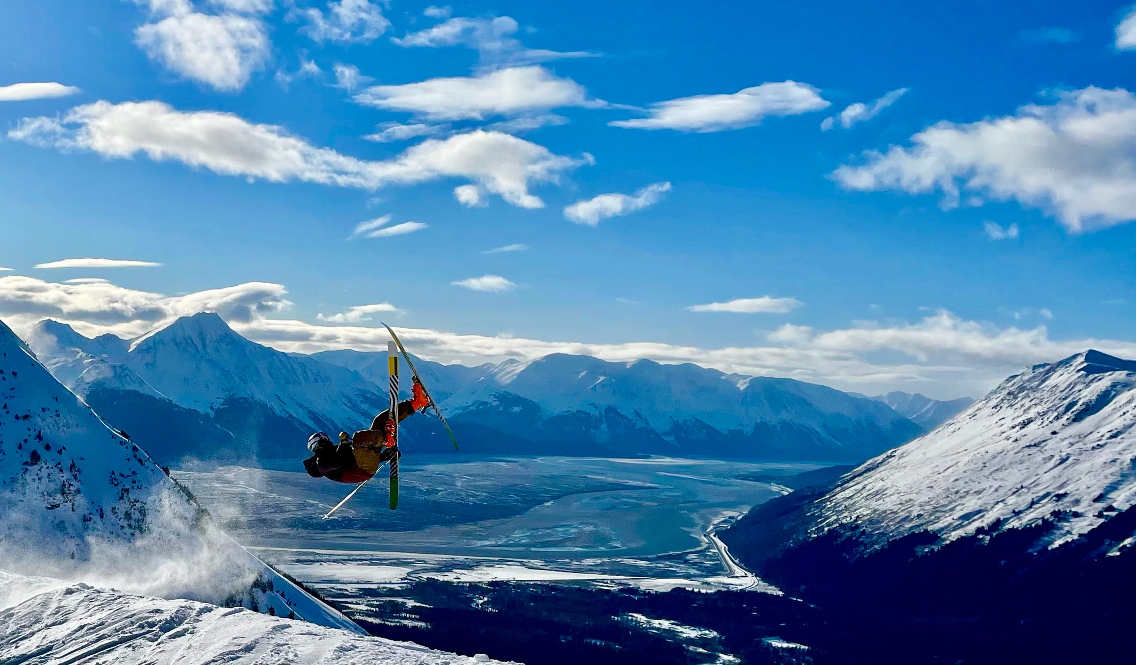 Skier launching upside down off a snowy ridge above a broad valley and snow-covered peaks in Alaska