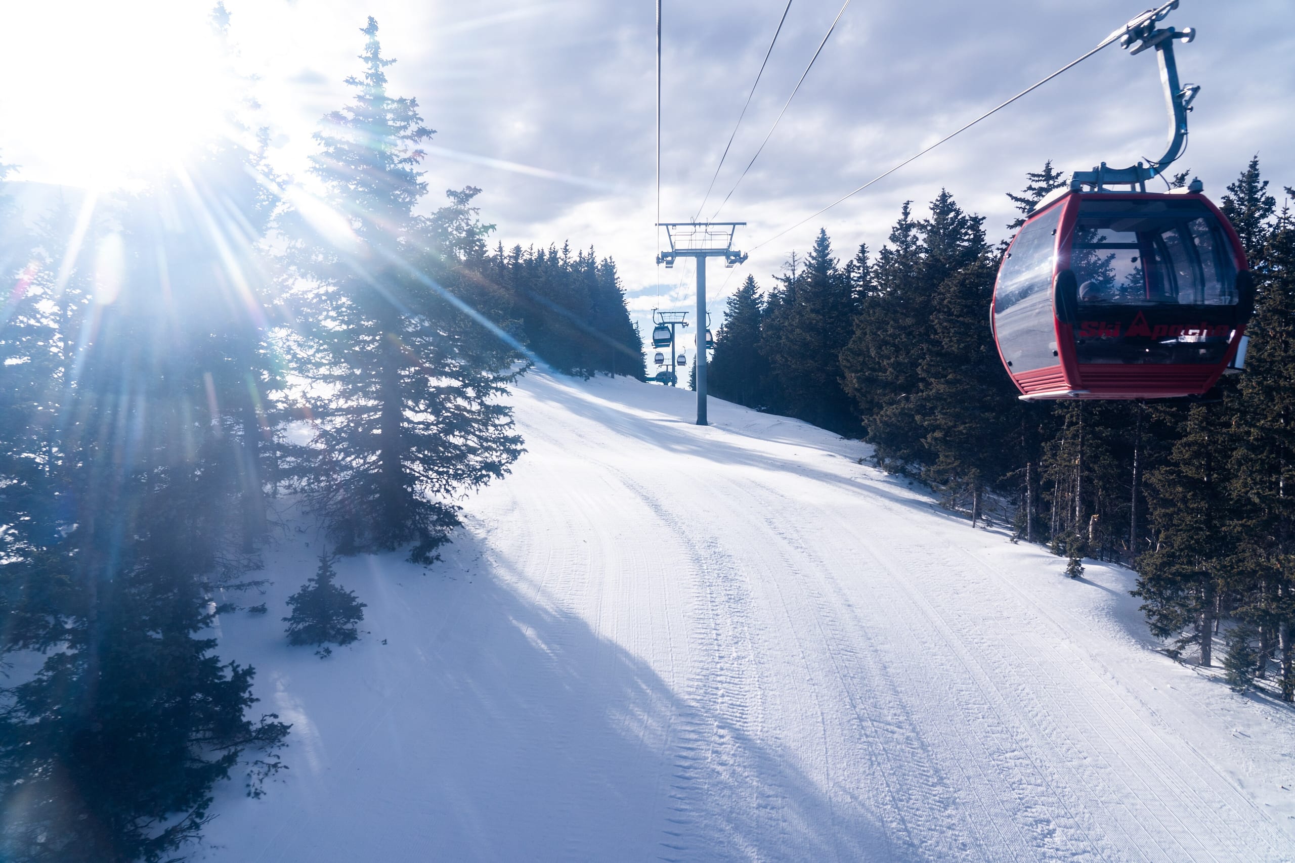 Red gondola cabin rising over a groomed snow run lined with conifers, sun flaring through trees