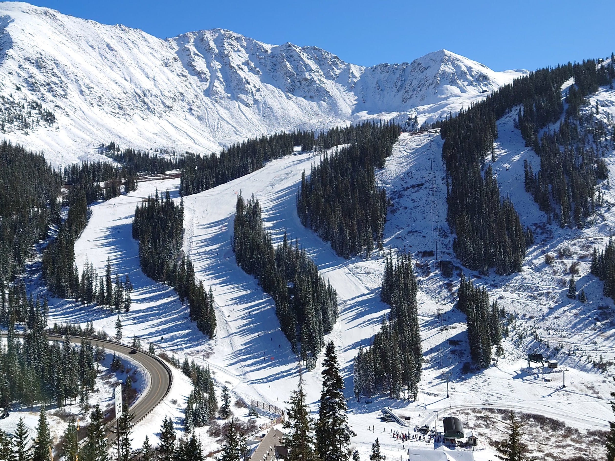 Snow-covered lower trails and base area of Arapahoe Basin below rocky ridges, with US 6 curving past