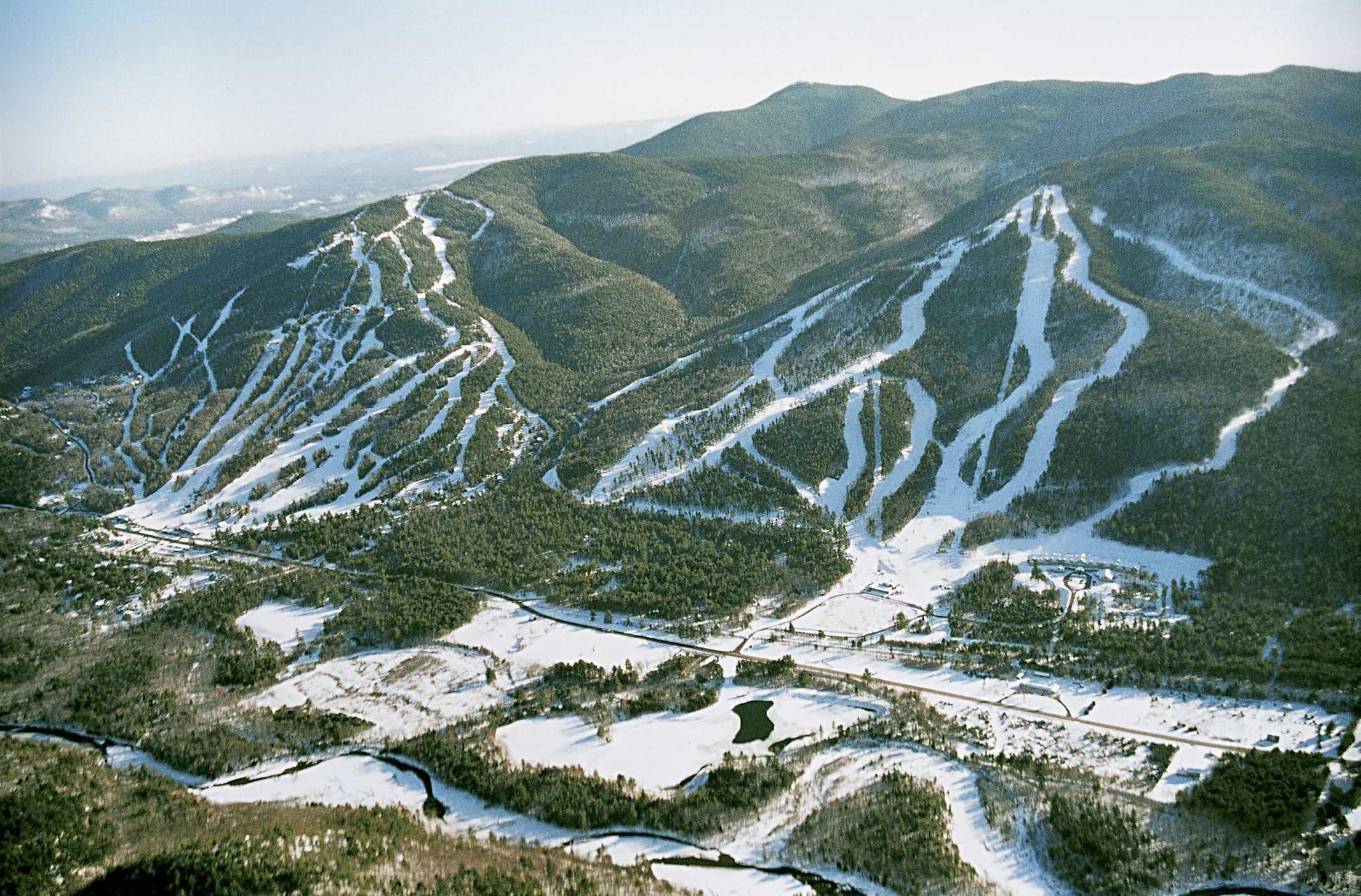 Aerial view of a two-peak ski area with snow-covered trails, base area and a highway in the valley