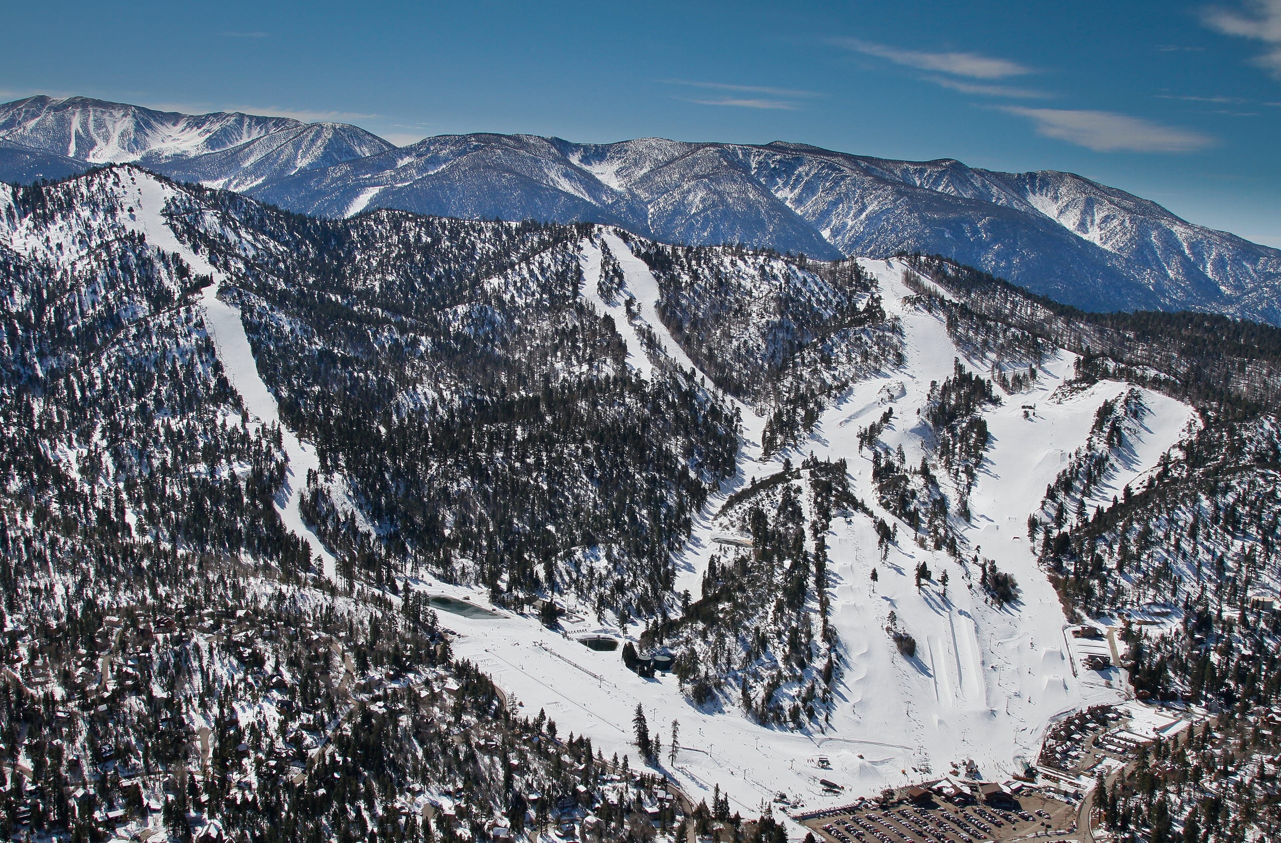 Aerial view of a snow-covered ski mountain with cut runs, lifts and a base area with parking lot below