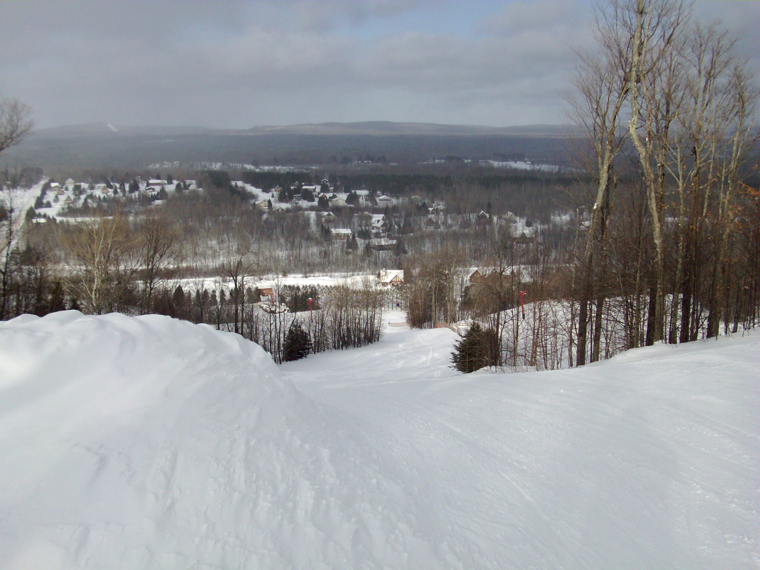 Snow-covered ski run looking down toward a small town and forested hills under grey skies