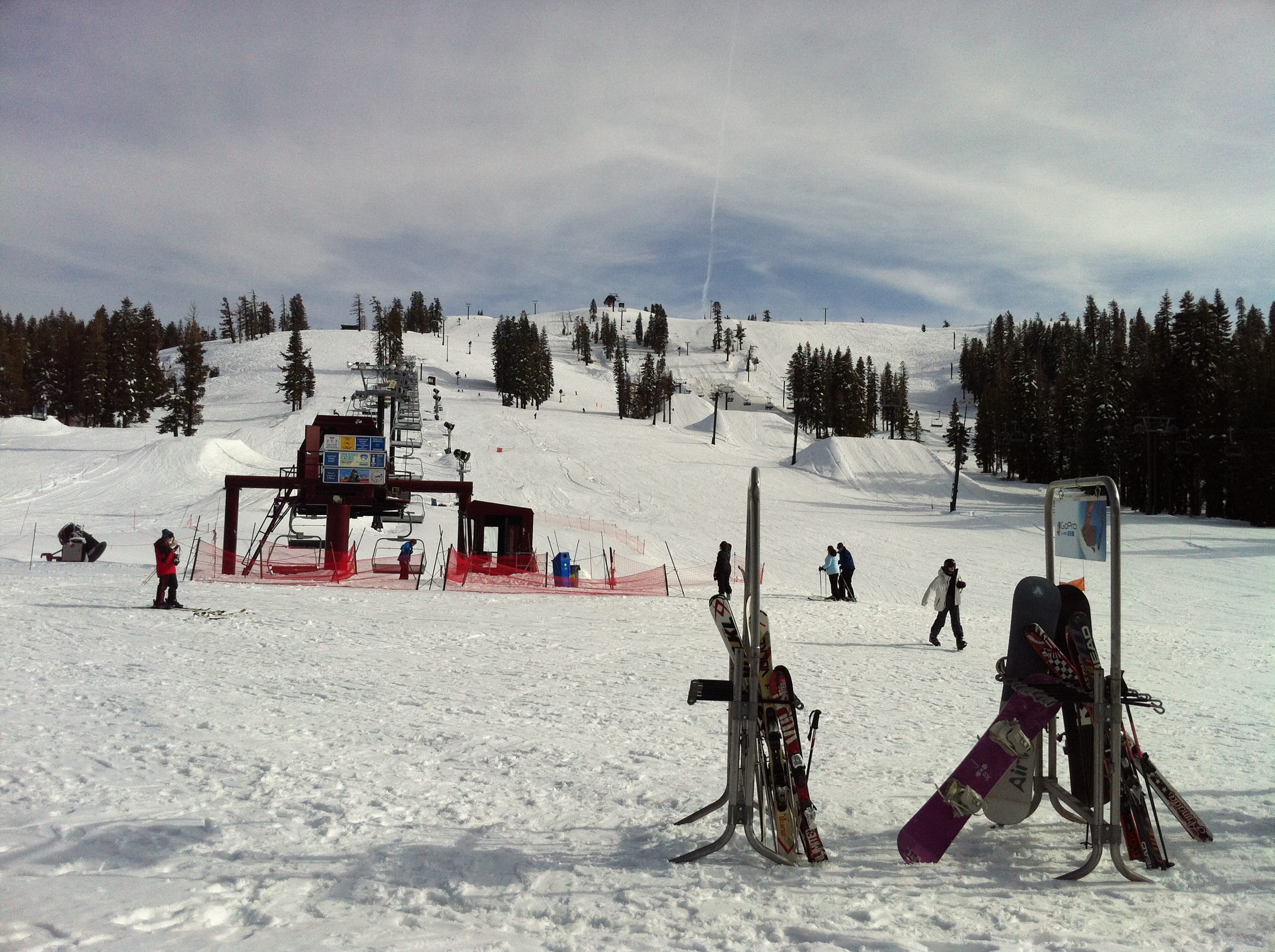 Snowy base area with a chairlift, ski racks and a few skiers below gentle tree-lined slopes