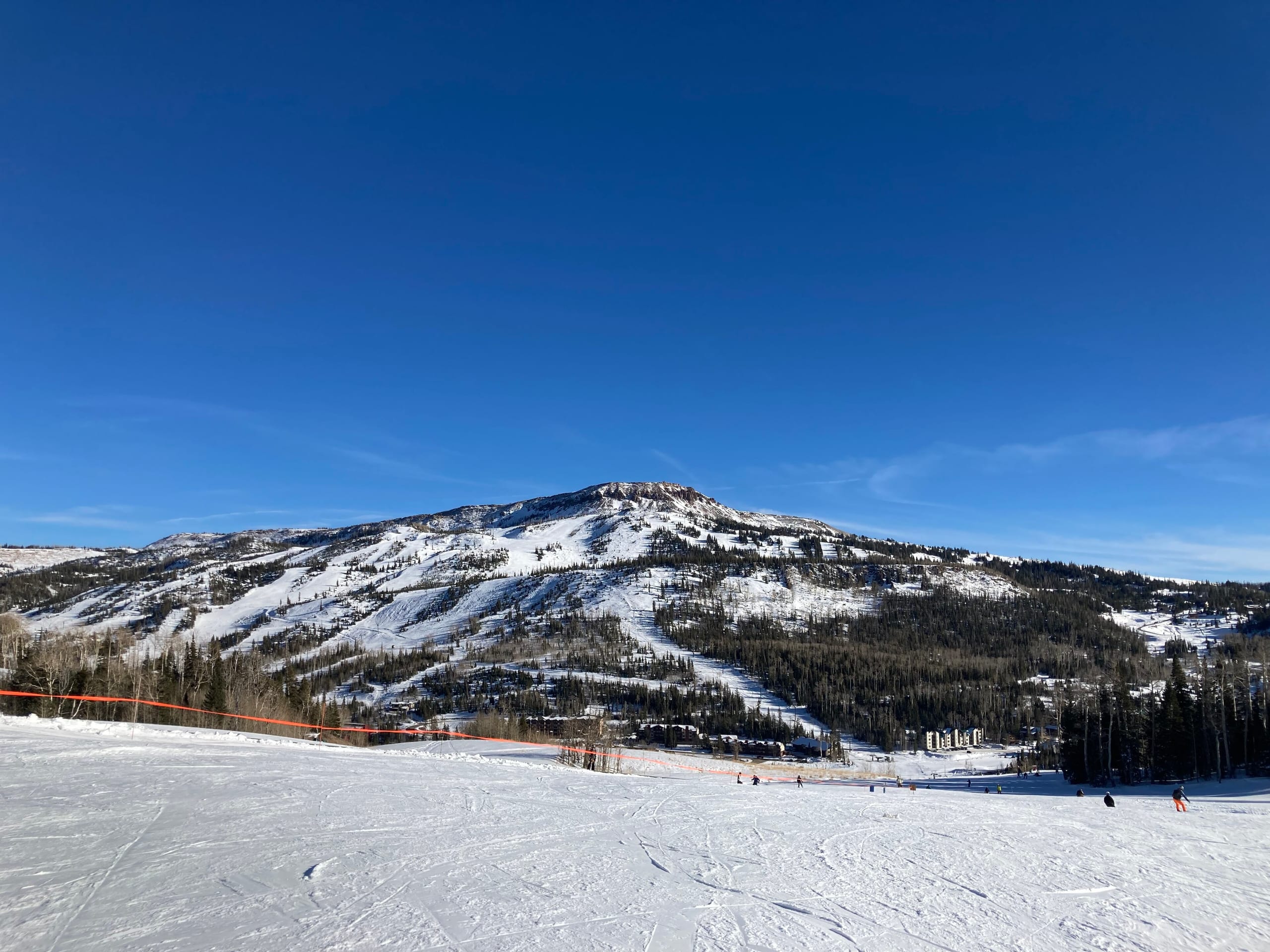 Snow-covered ski runs and forested slopes of Brian Head Peak under a clear blue sky, with skiers on a groomed run in the foreground