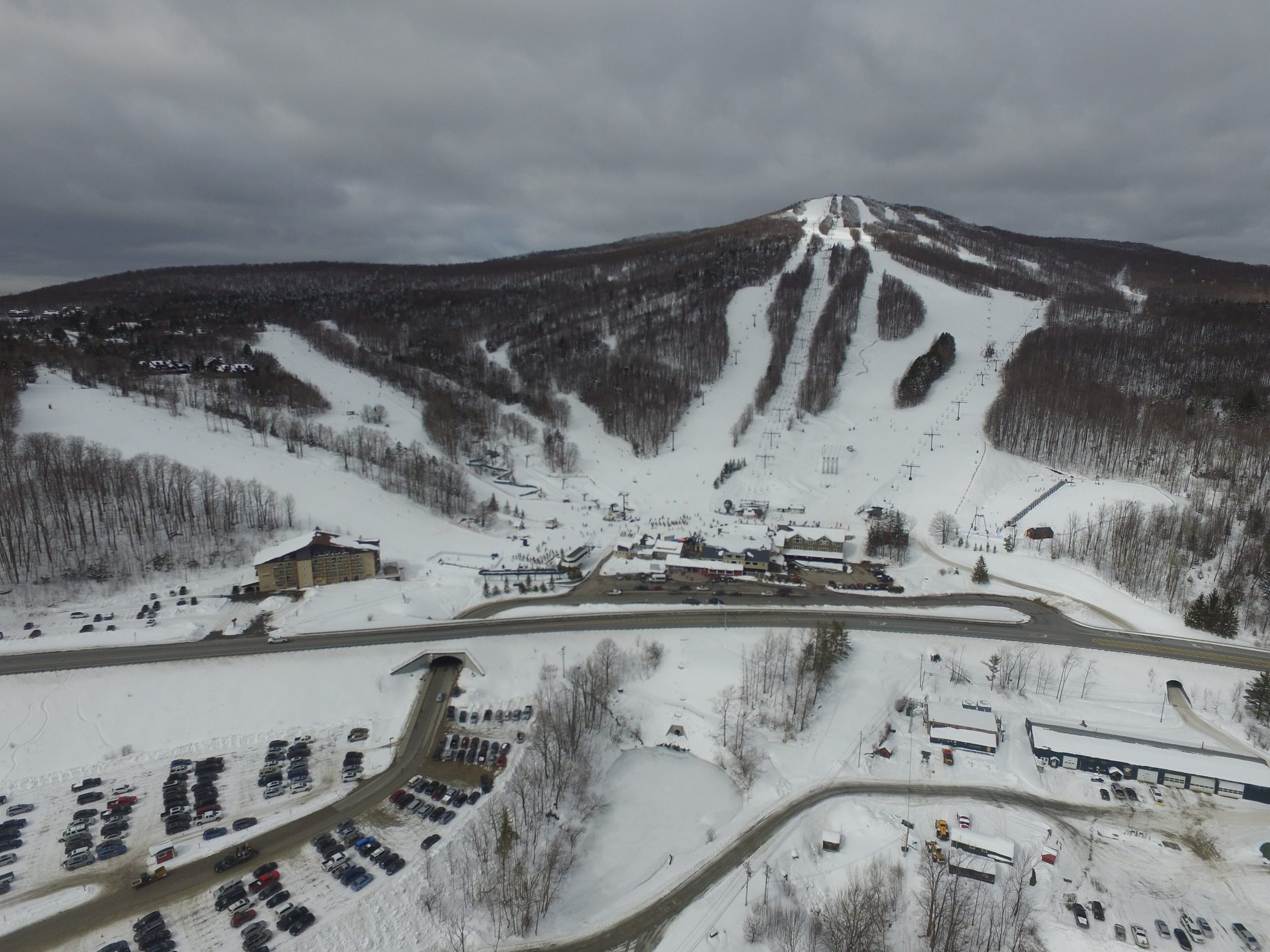 Aerial view of a snow-covered ski mountain with trails, base lodge, parking lots and highway below