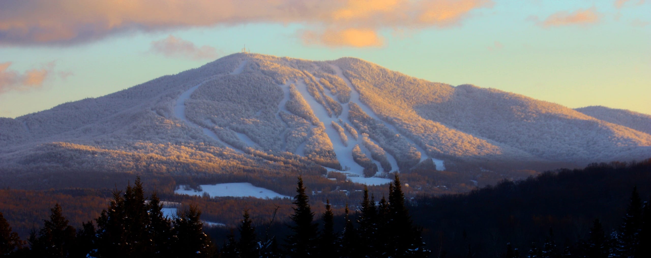 Snow-covered Burke Mountain at sunrise with ski trails cut down its face, seen from across a forested valley