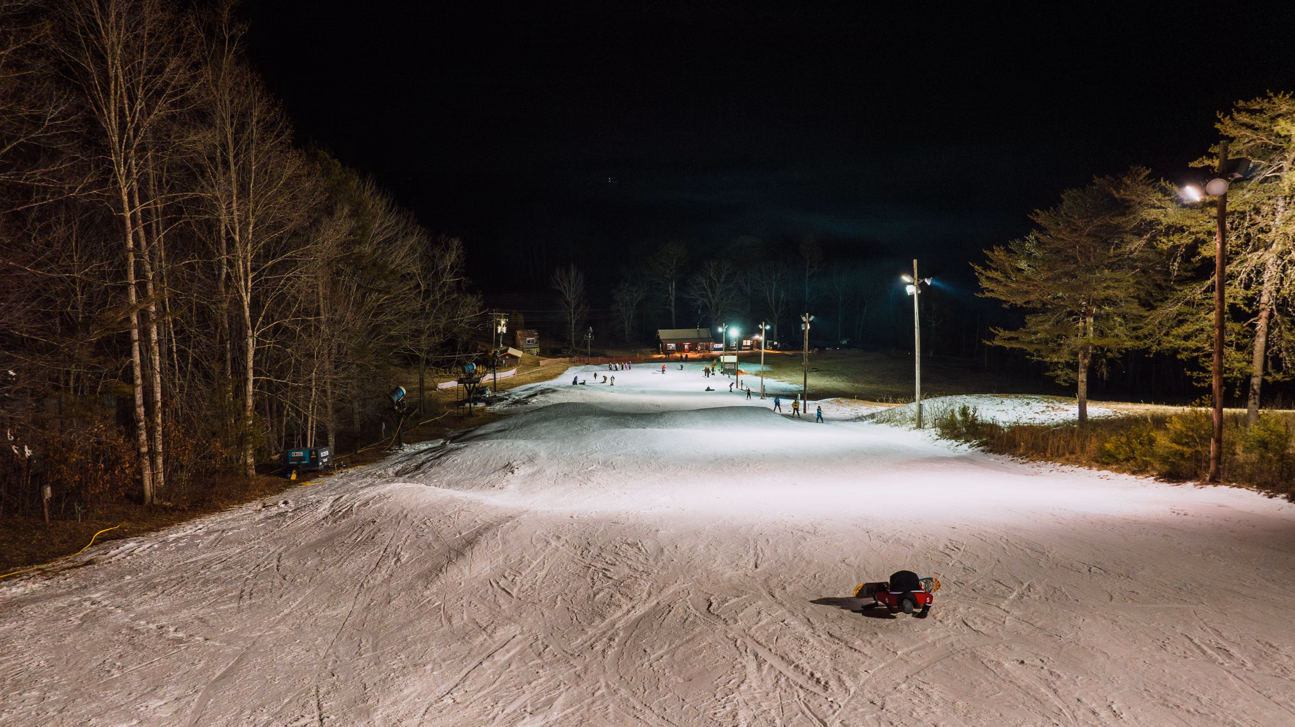 Floodlit ski slope at night, small figures and a lit lodge at the top of the run.