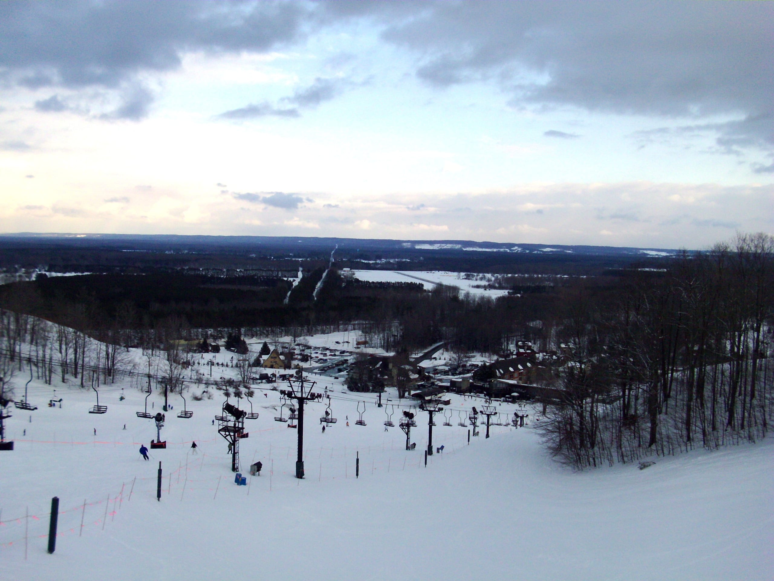 Snow-covered ski slope with chairlift descending to a base village under a cloudy sky