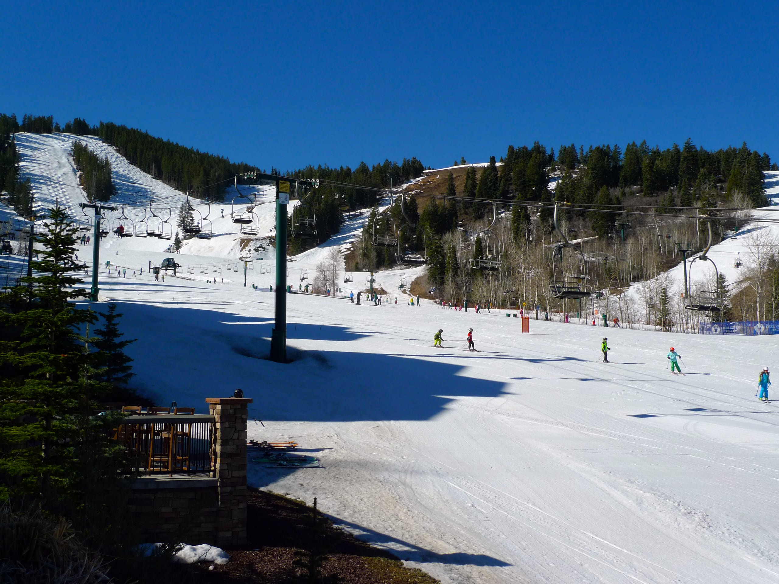 Skiers on a wide groomed run below chairlifts and forested slopes under a clear blue sky