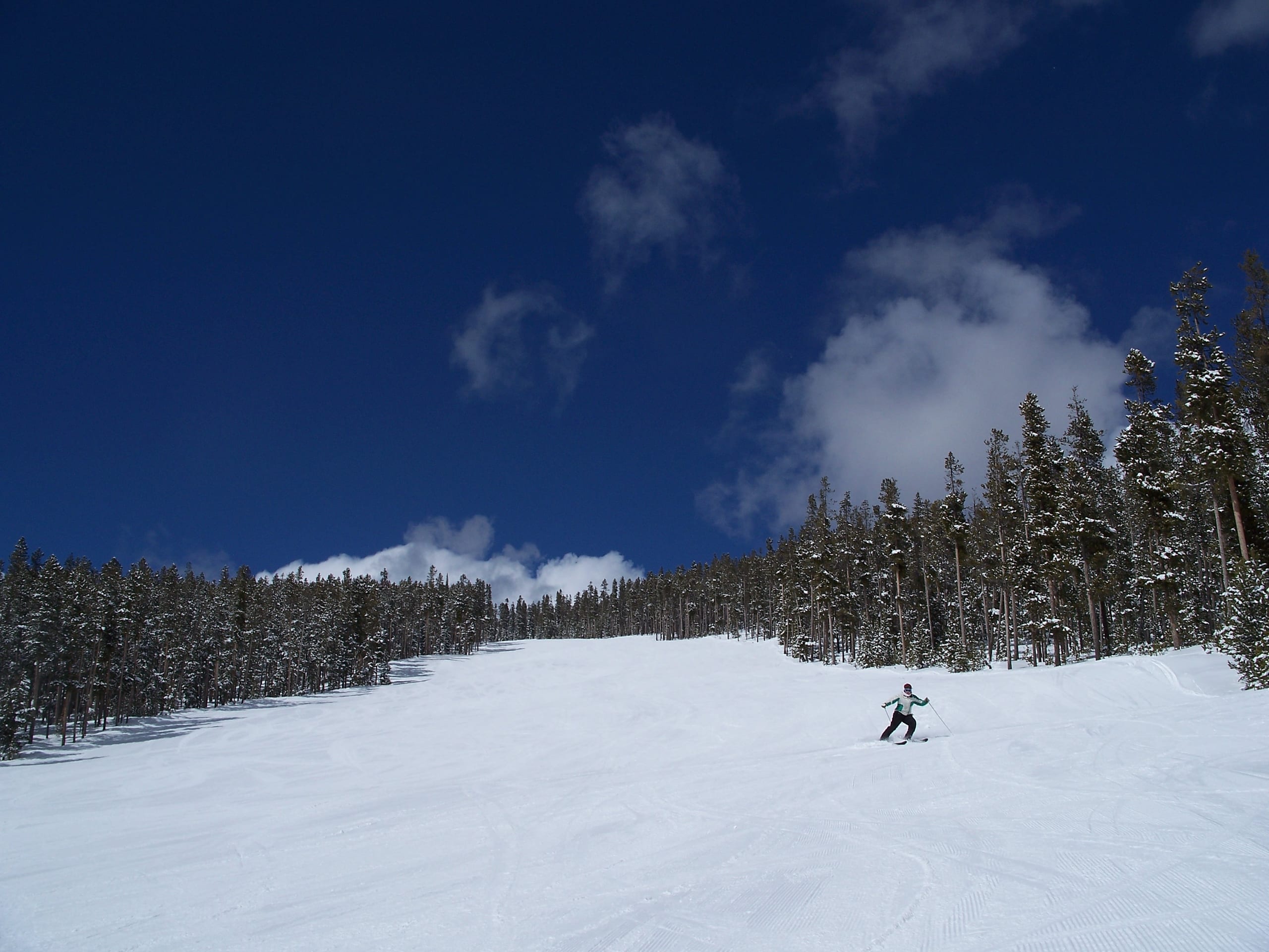 A lone skier descends a wide groomed run bordered by snow-covered conifers under a deep blue sky.