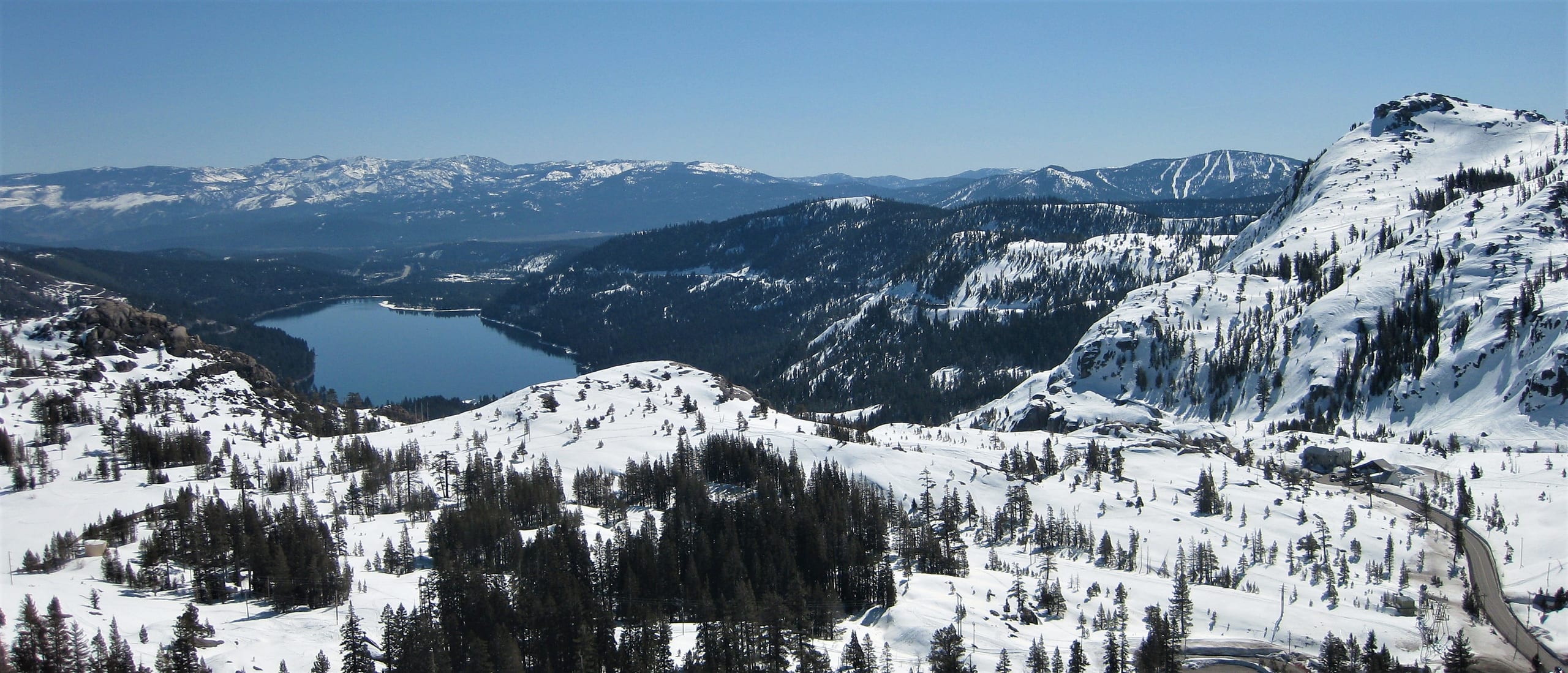 Snowy view east from Donner Pass over Donner Lake, with Donner Peak and forested ridges under blue sky