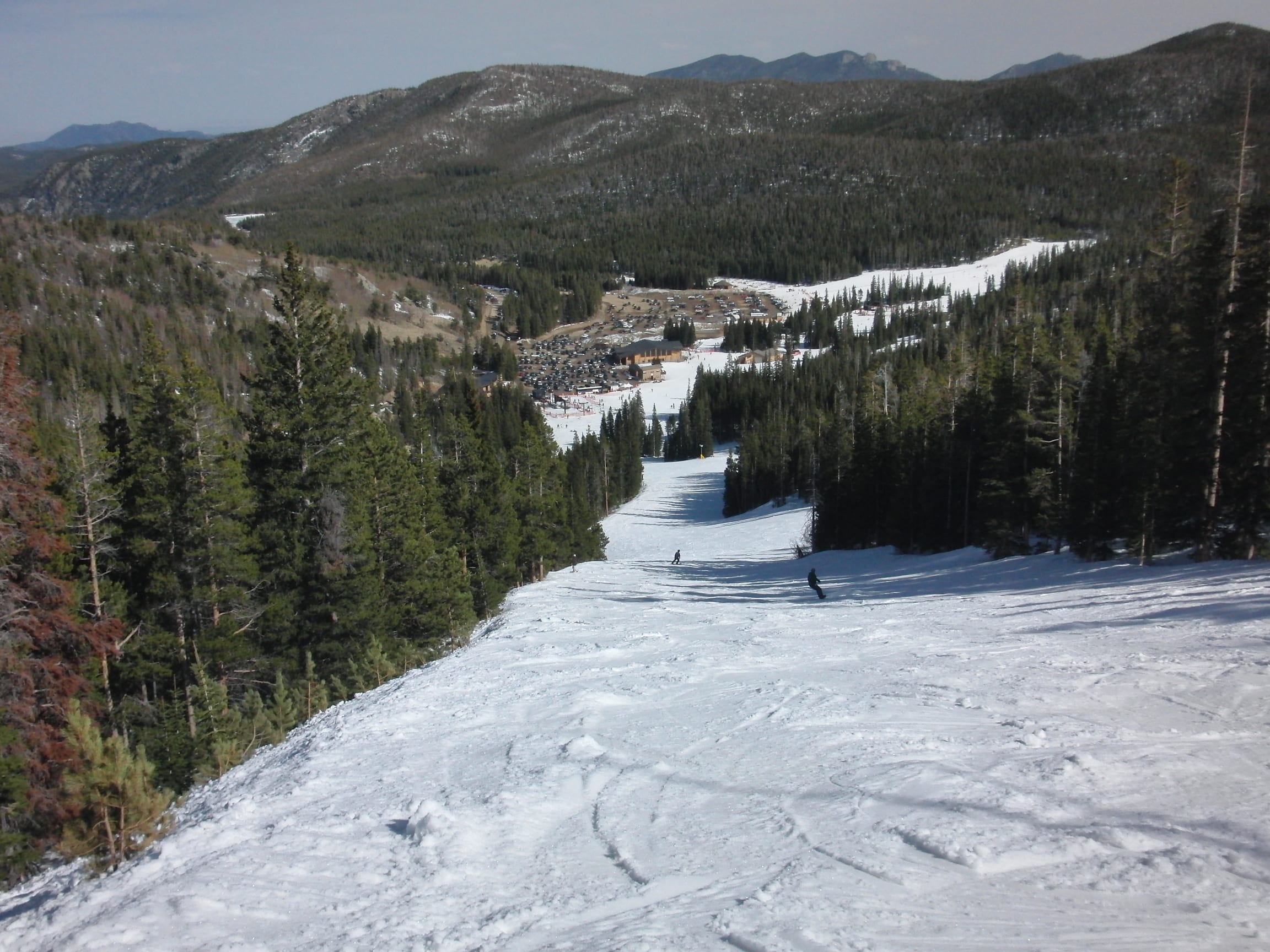 View down a snowy ski run toward Eldora's base lodge, with two skiers and forested ridges beyond