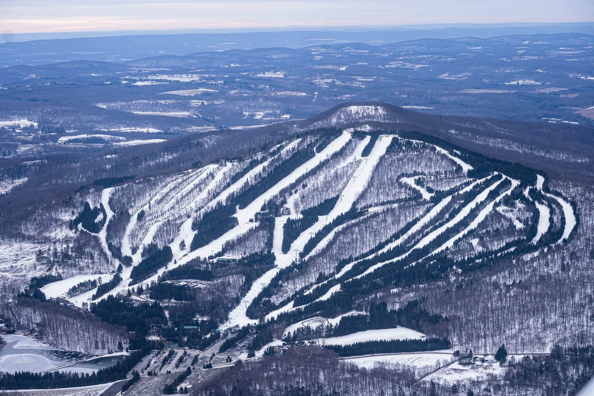 Aerial view of a ski mountain with snow-covered trails fanning down to a base area and parking lot