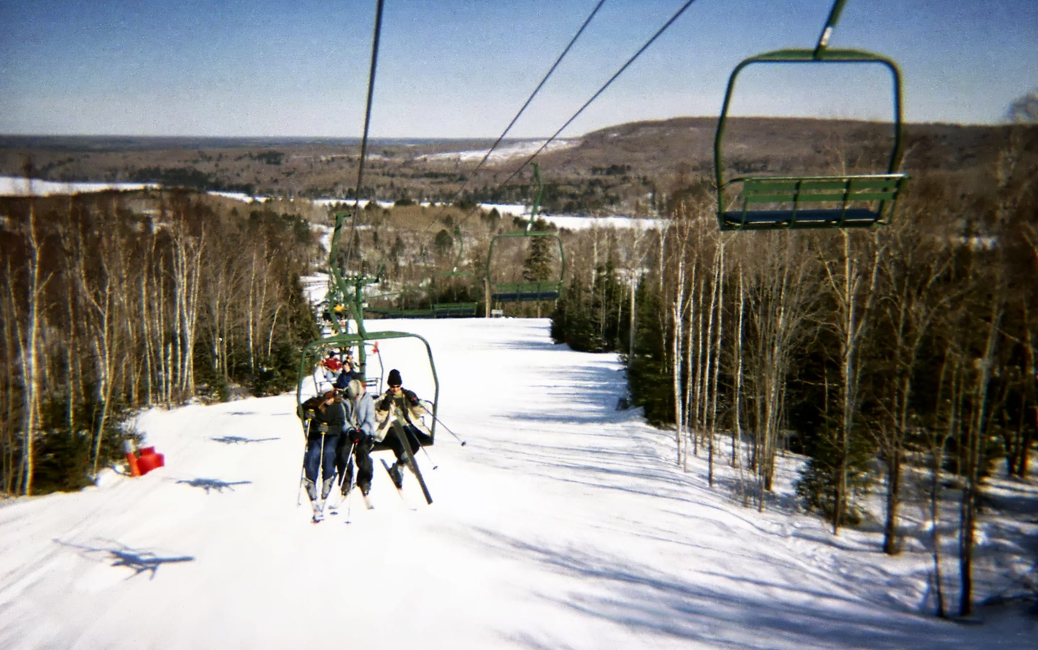 Skiers ride a chairlift up a snow-covered run lined with bare trees, with a wooded valley beyond