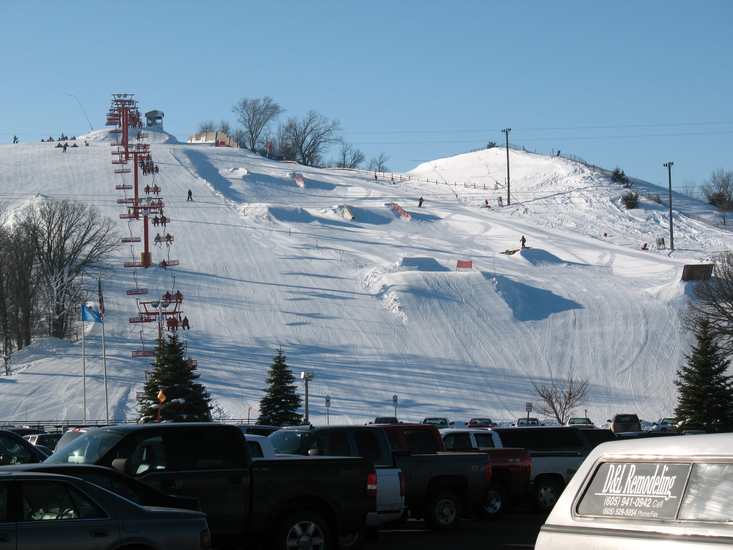 Snow-covered ski hill with chairlift and terrain park jumps above a parking lot full of vehicles