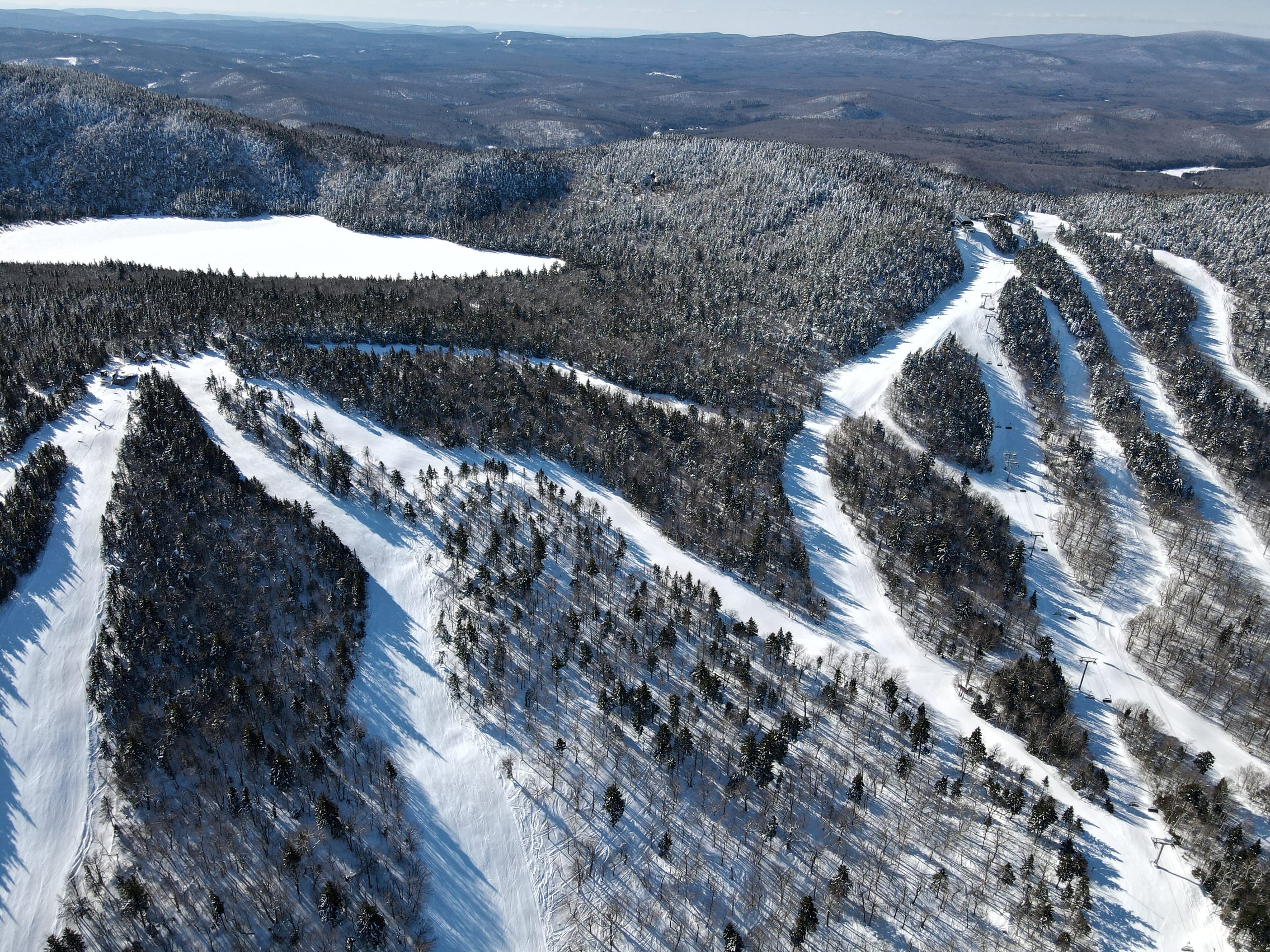 Aerial view of snow-covered ski trails and glades on Haystack Mountain with a frozen pond and forested hills beyond