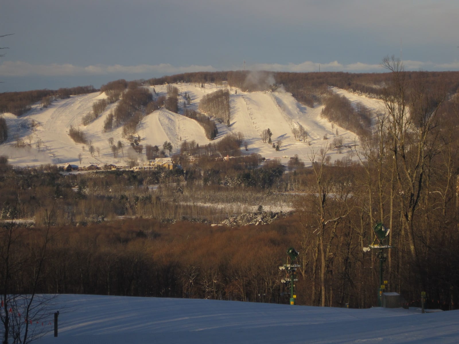 Snow-covered ski runs and lifts on a wooded hill with base lodges below, seen from an opposing slope