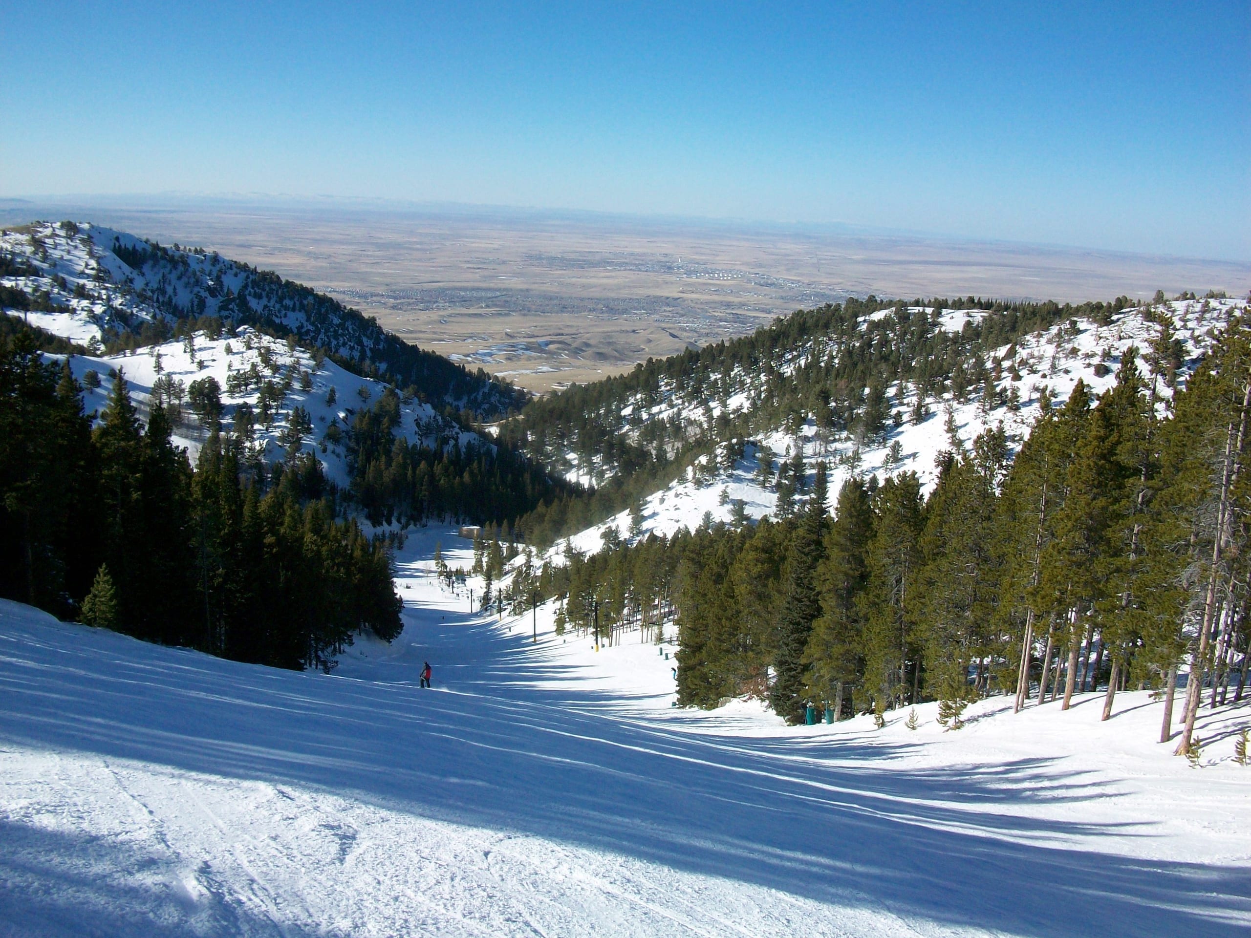 Snow-covered ski run descending through pines with the plains near Casper, Wyoming visible below
