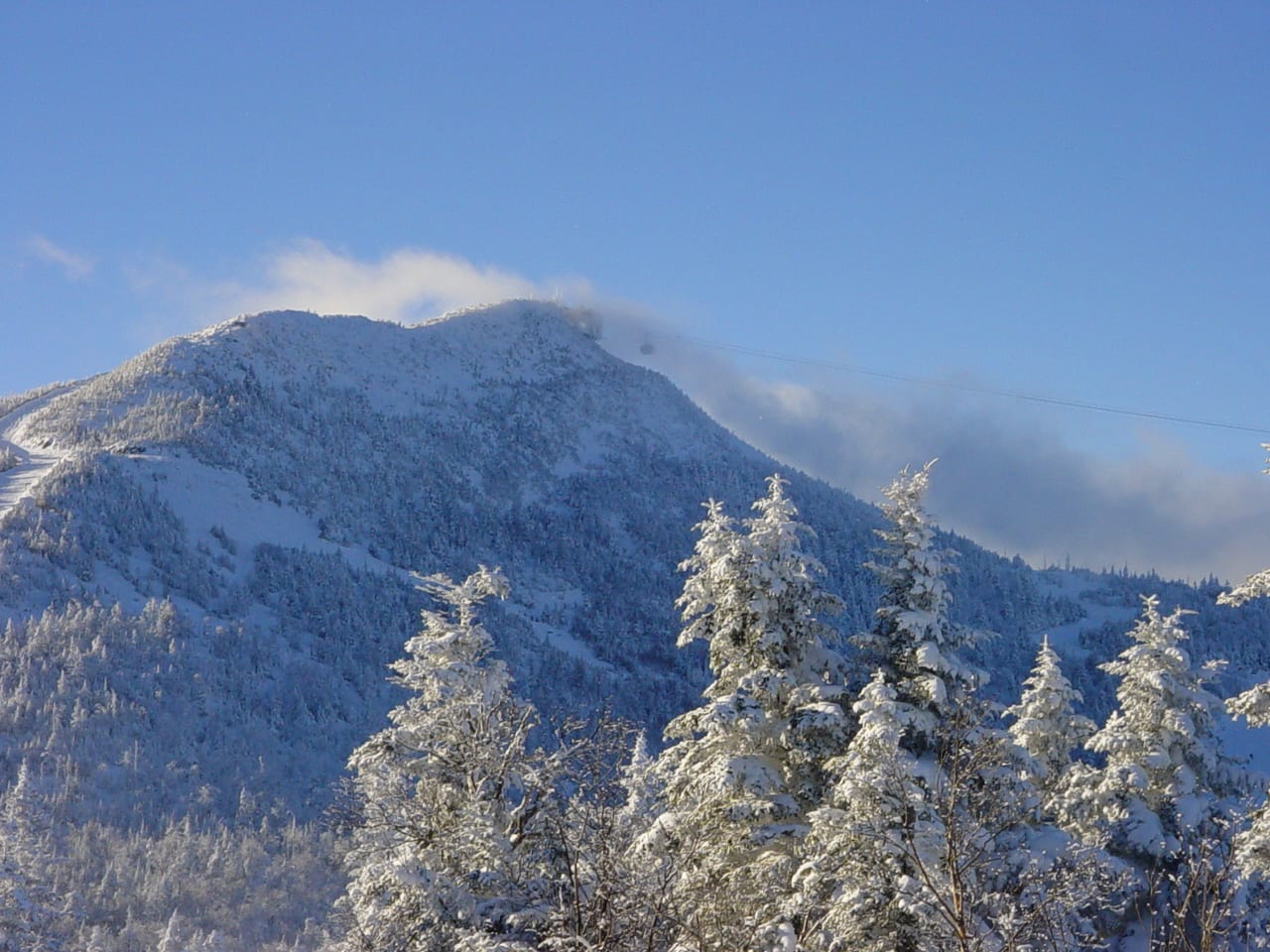 Snow-covered summit of Jay Peak under blue sky, with a tramway cable and rimed spruce in front.