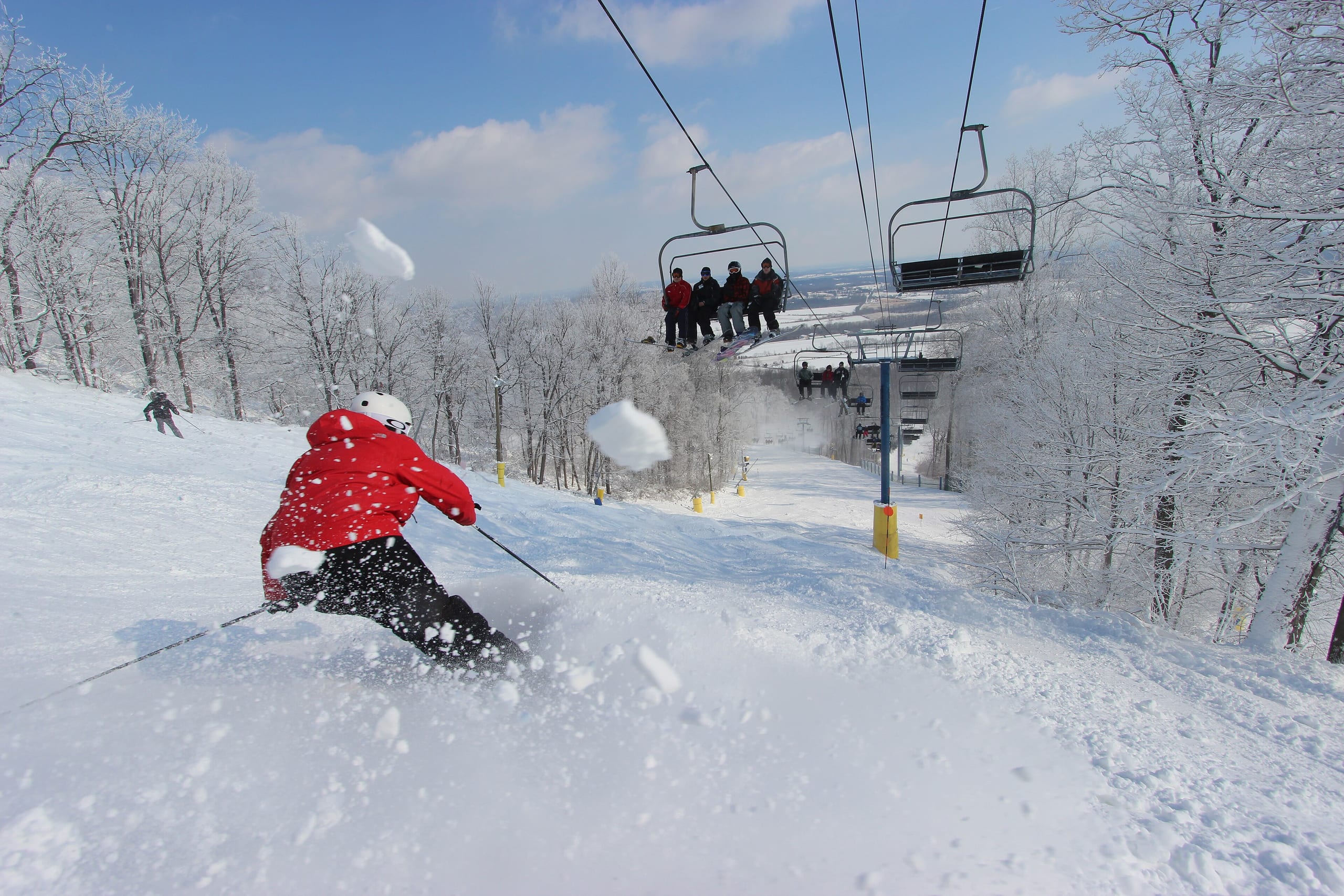 A skier in a red jacket carves down a snowy run beneath a chairlift with riders overhead