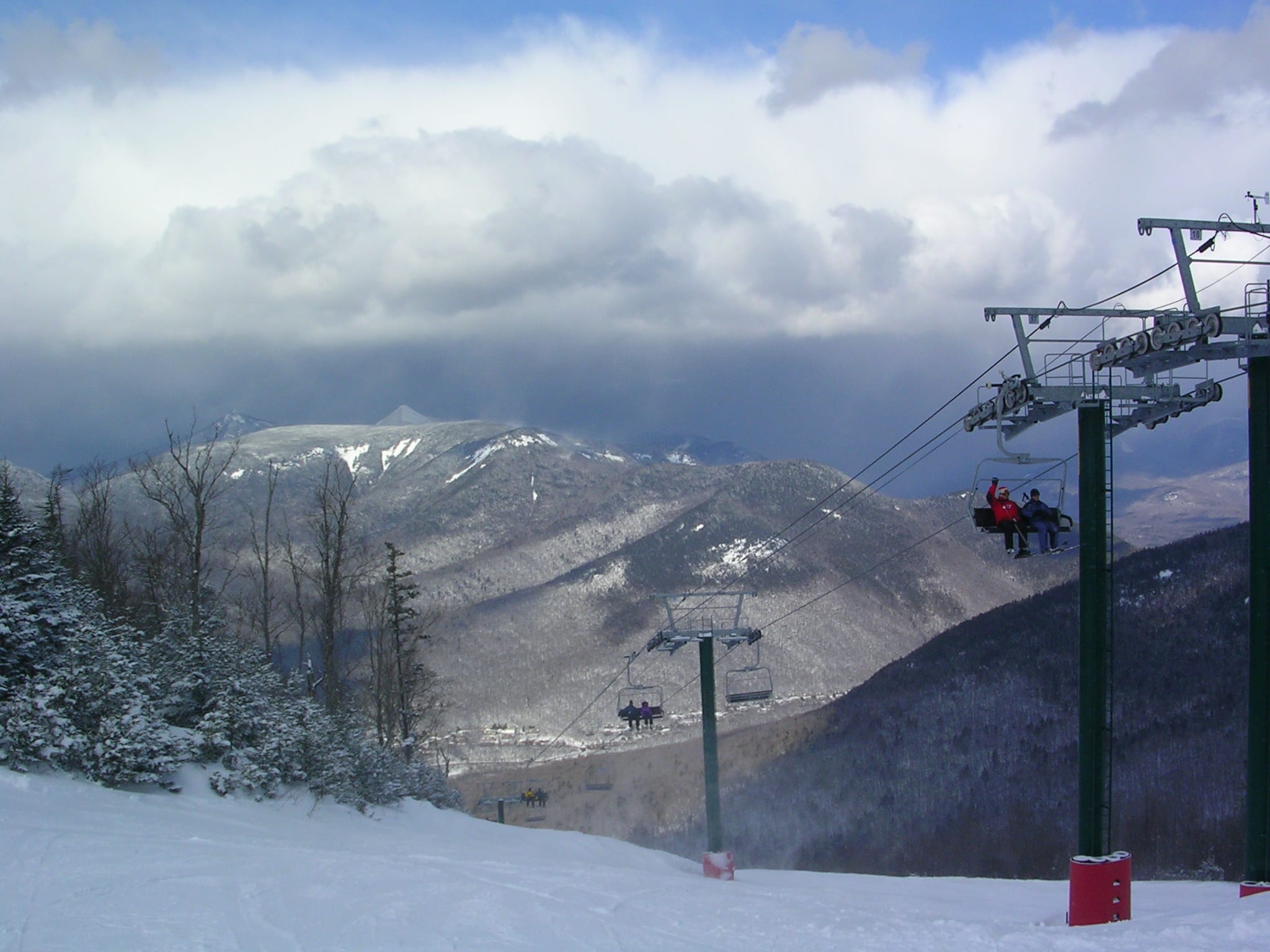 Skiers on a chairlift above a snowy trail at Loon Mountain, with cloud-topped peaks across the valley