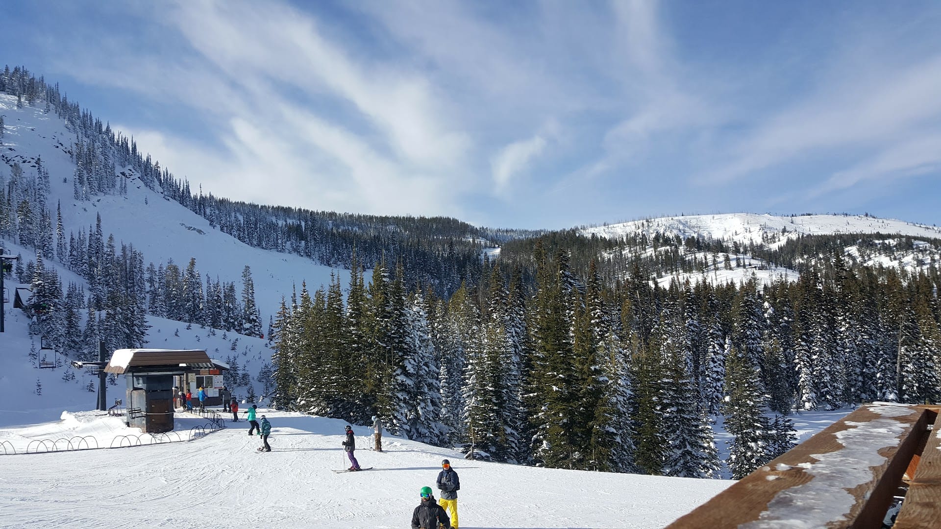 Skiers on a snowy base slope below a chairlift terminal, with forested snow-covered ridges beyond