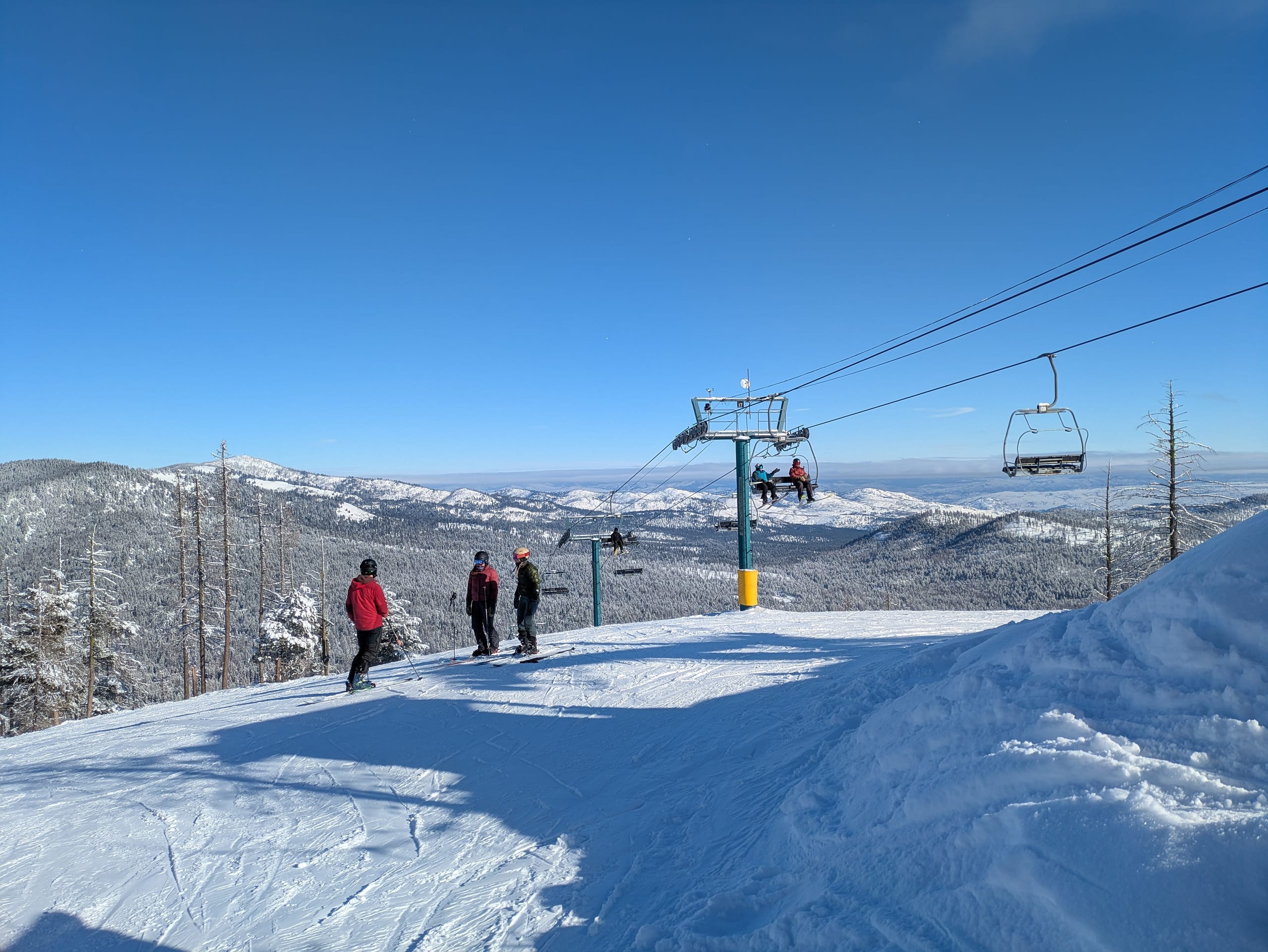 Skiers atop a groomed run beside a chairlift, with forested snowy ridges and a valley beyond
