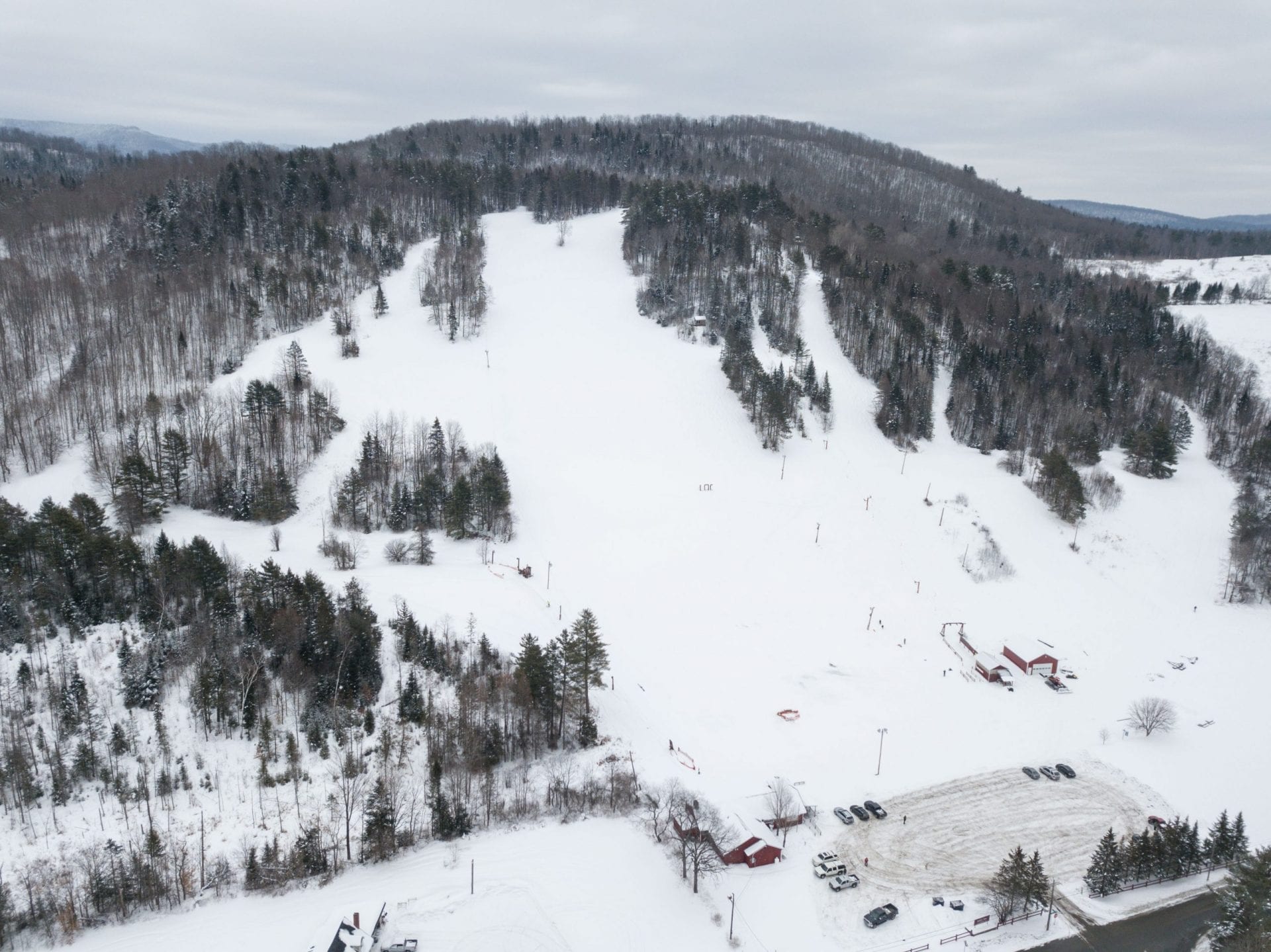Aerial view of a small snow-covered ski hill with cleared trails, a lift line, base lodge and parking lot