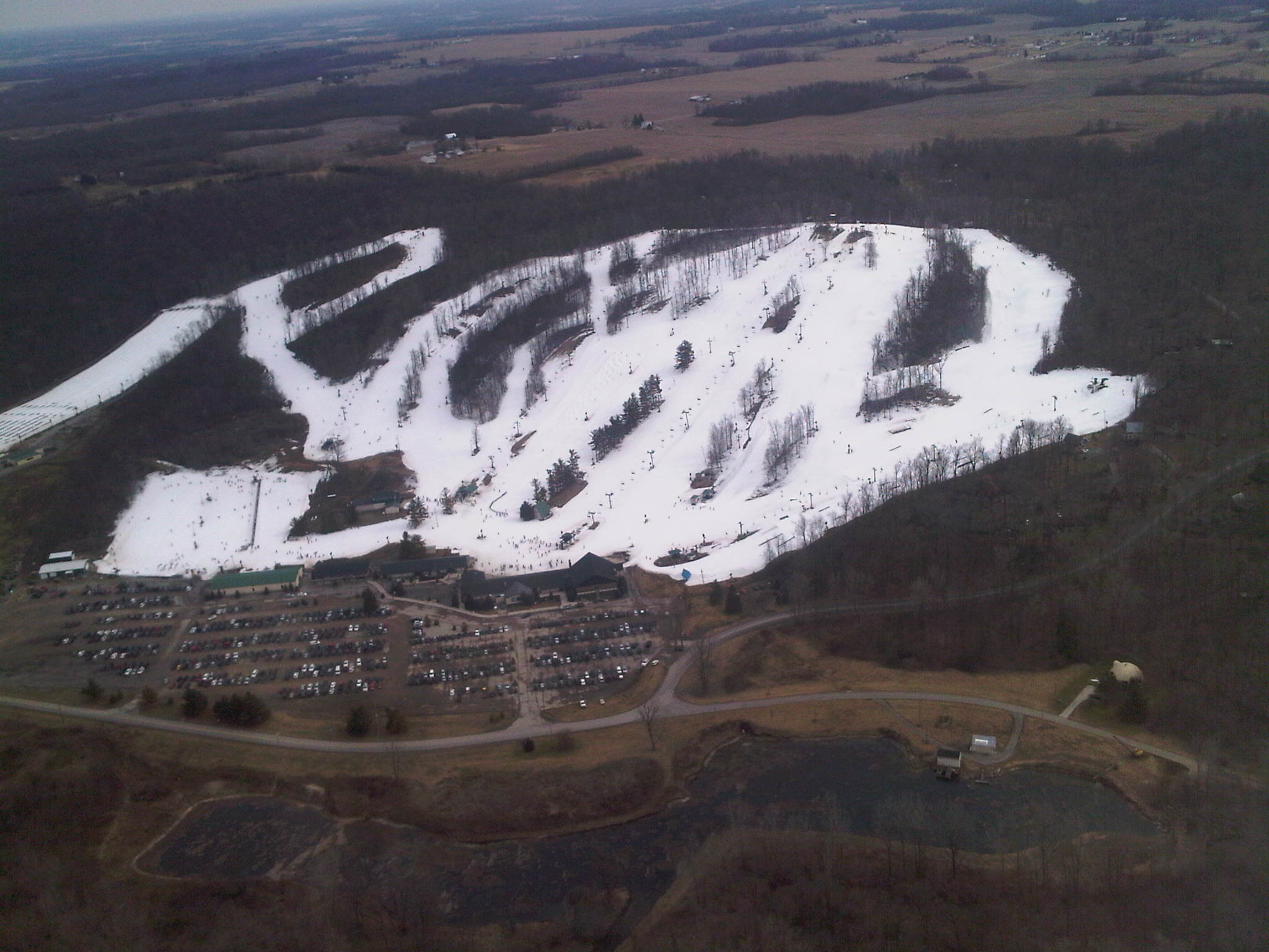 Aerial view of a small ski hill with white runs, lifts and a large parking lot amid brown fields