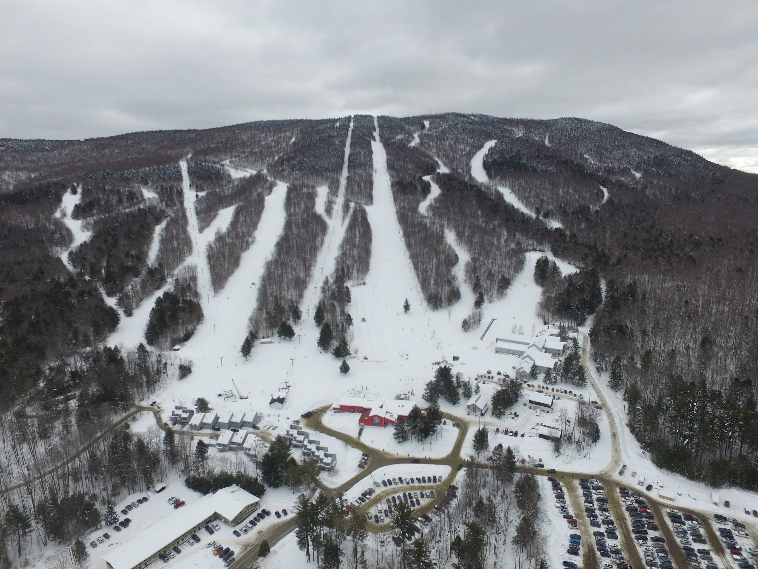 Aerial view of a ski area's snow-covered trails, base lodge and parking lots below a wooded ridge