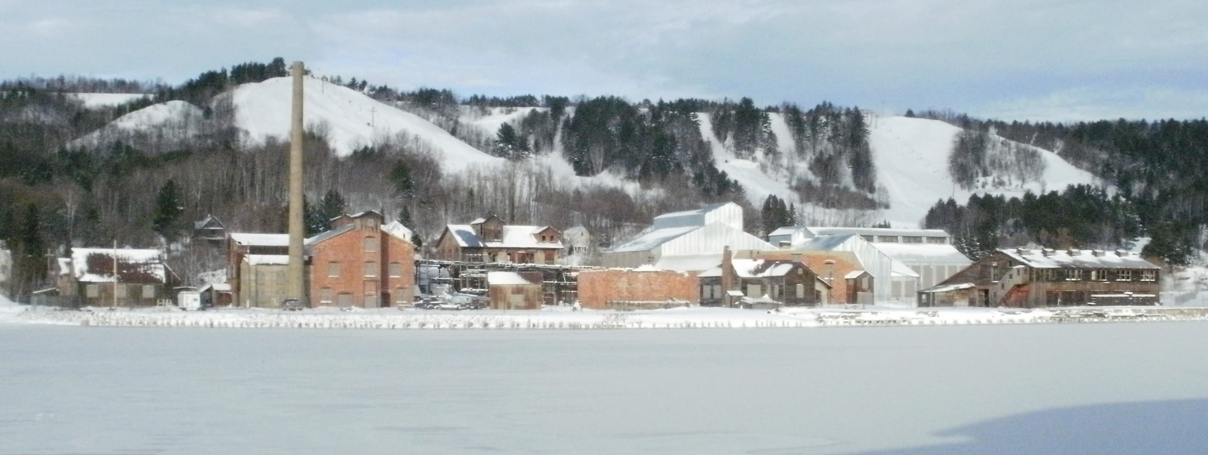 Snow-covered ski runs on a hill above the Quincy Smelter buildings and a frozen waterway in winter