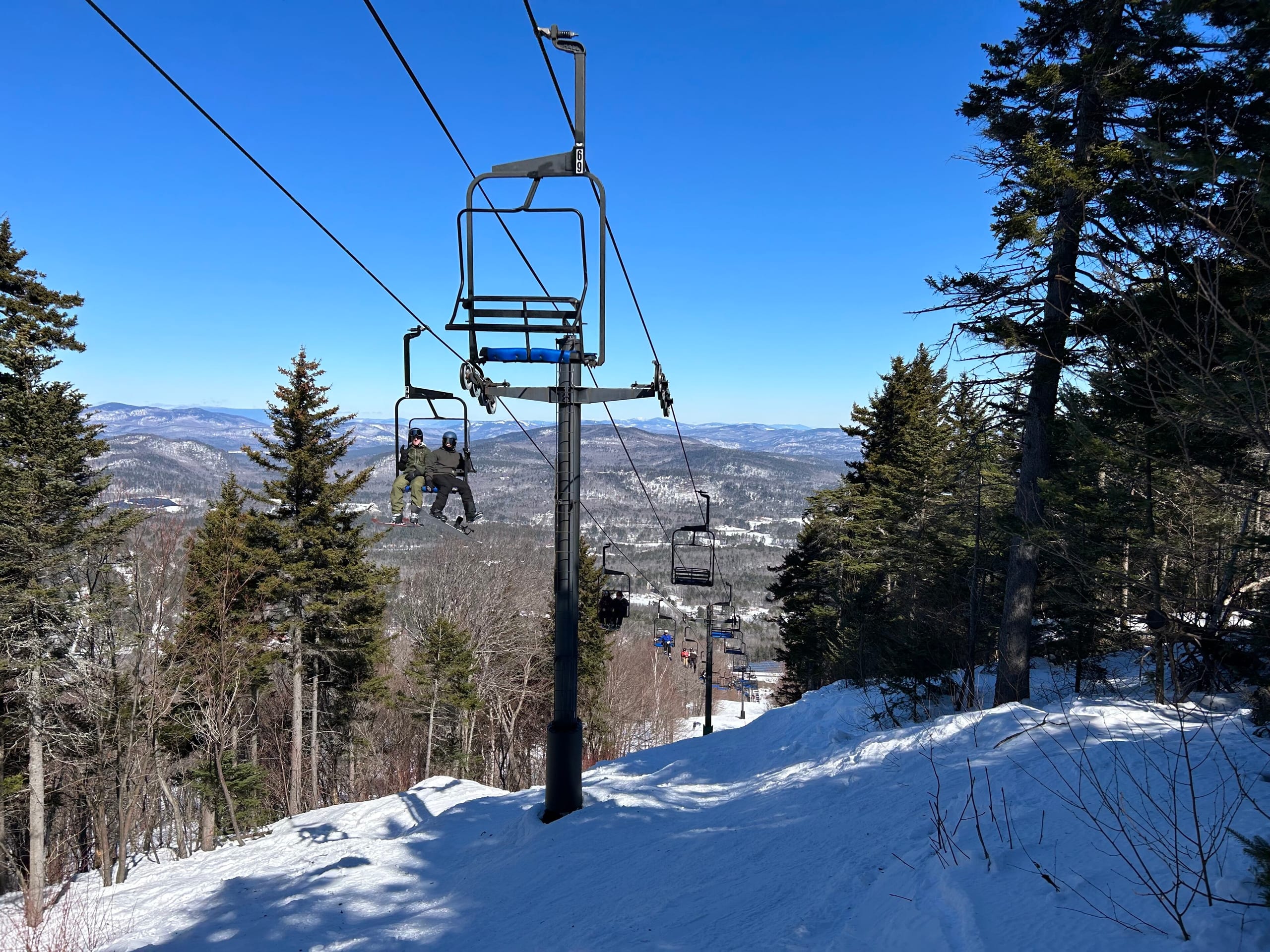 Skiers ride a chairlift above a snowy trail with forested hills in the distance
