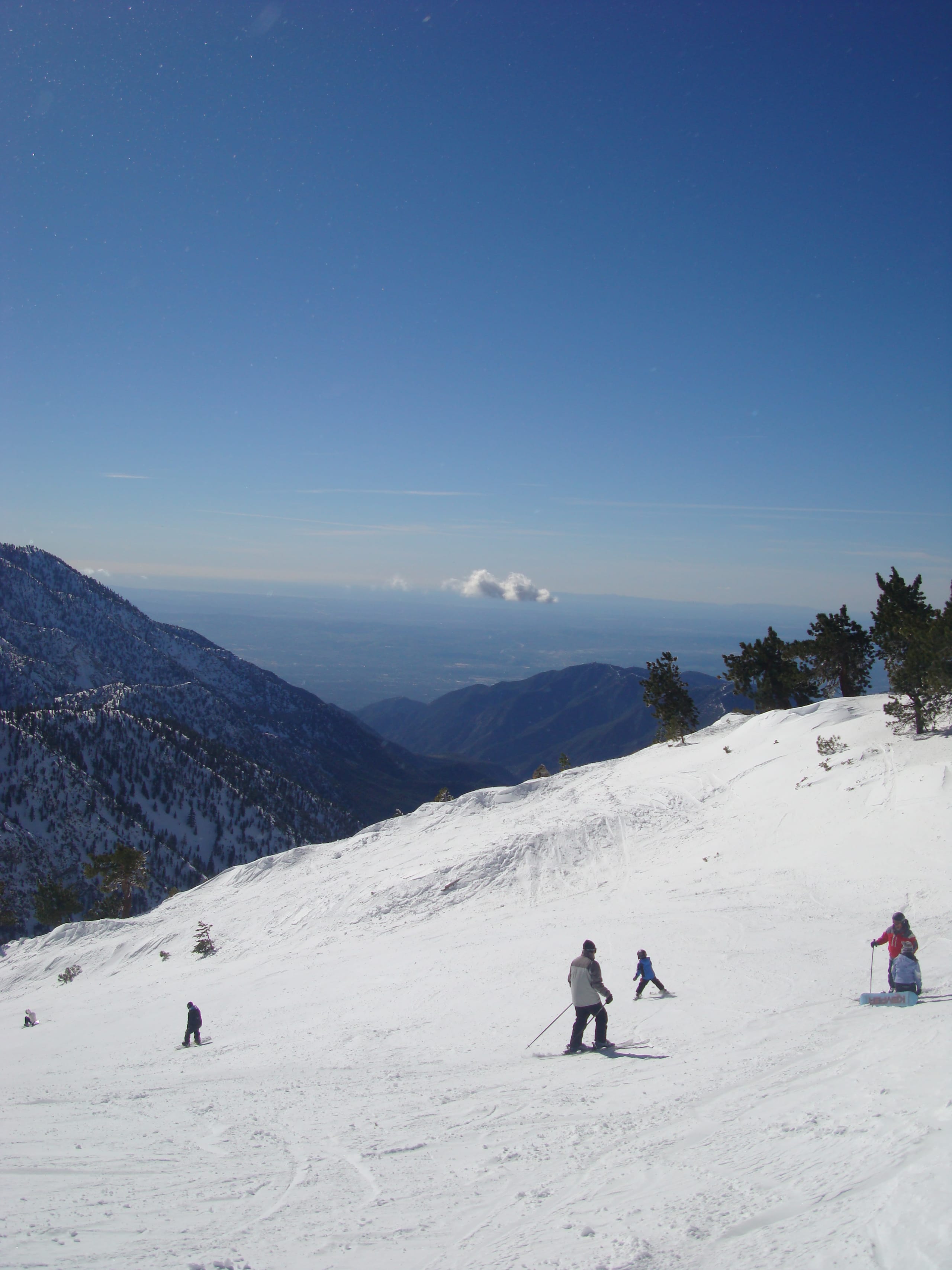 Skiers descend a wide snowy run at Mount Baldy above forested ridges and a hazy valley below.