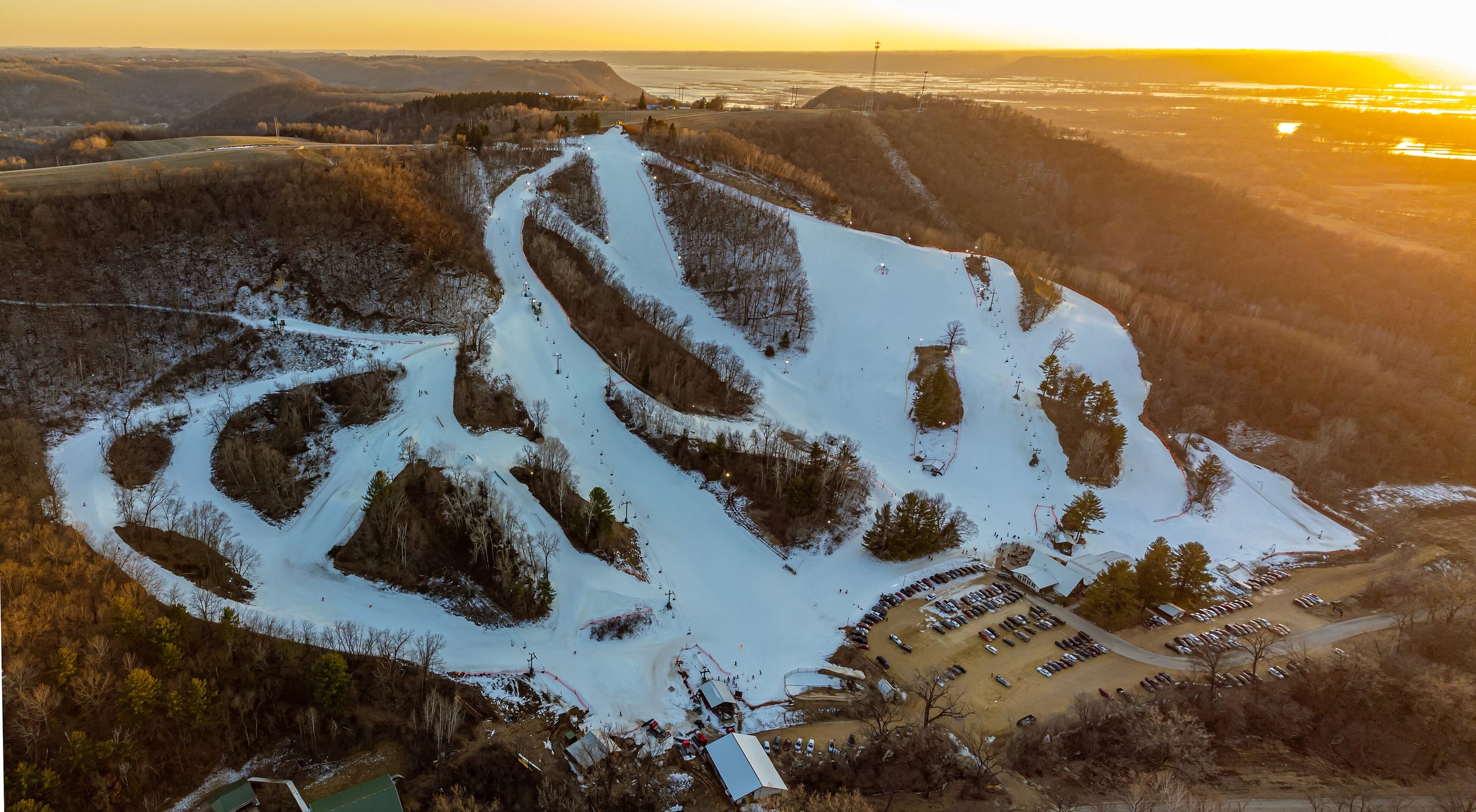 Aerial view at sunset of snow-covered ski runs, lifts and base parking lots on a bluff above a river valley