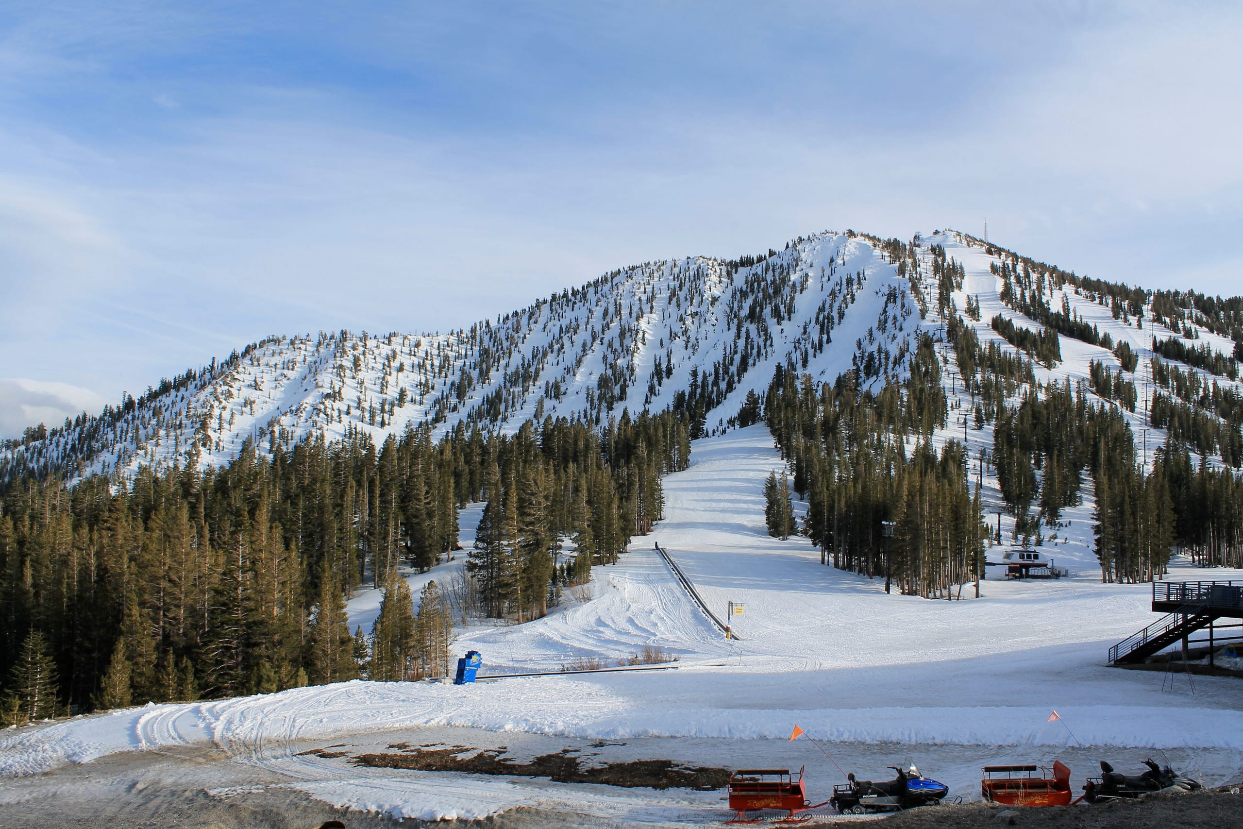 Snow-covered ski runs and forested slopes rising above a base area with snowmobiles and a lift terminal