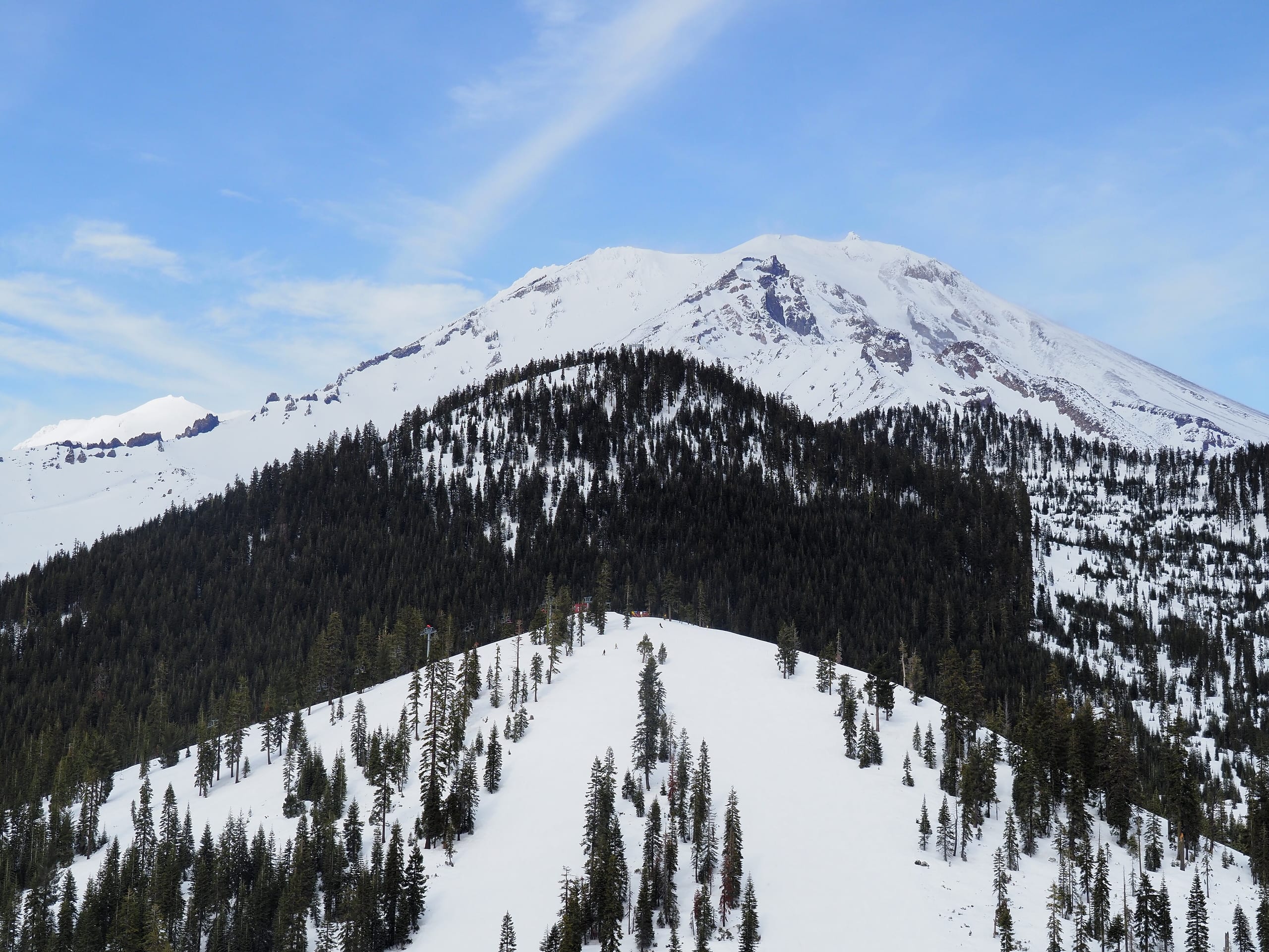 Snow-covered Mount Shasta rising behind a forested ridge with ski runs and a lift line below