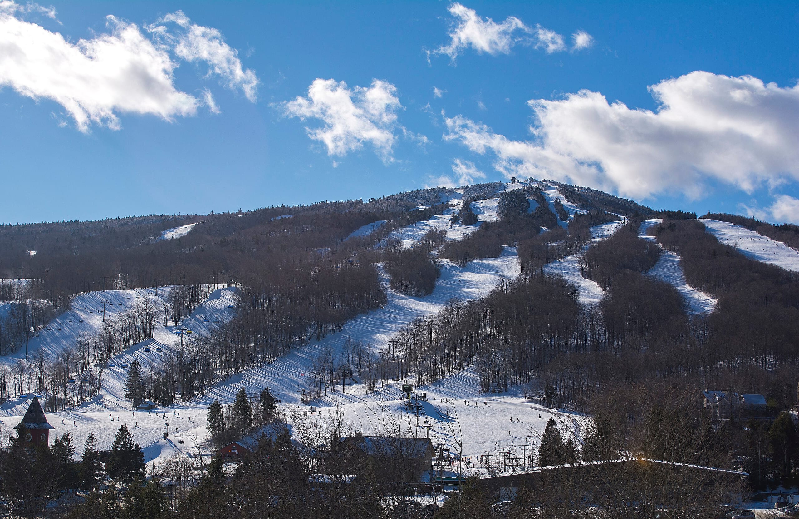 Snow-covered trails and lifts fan down the broad face of Mount Snow under blue sky with clouds