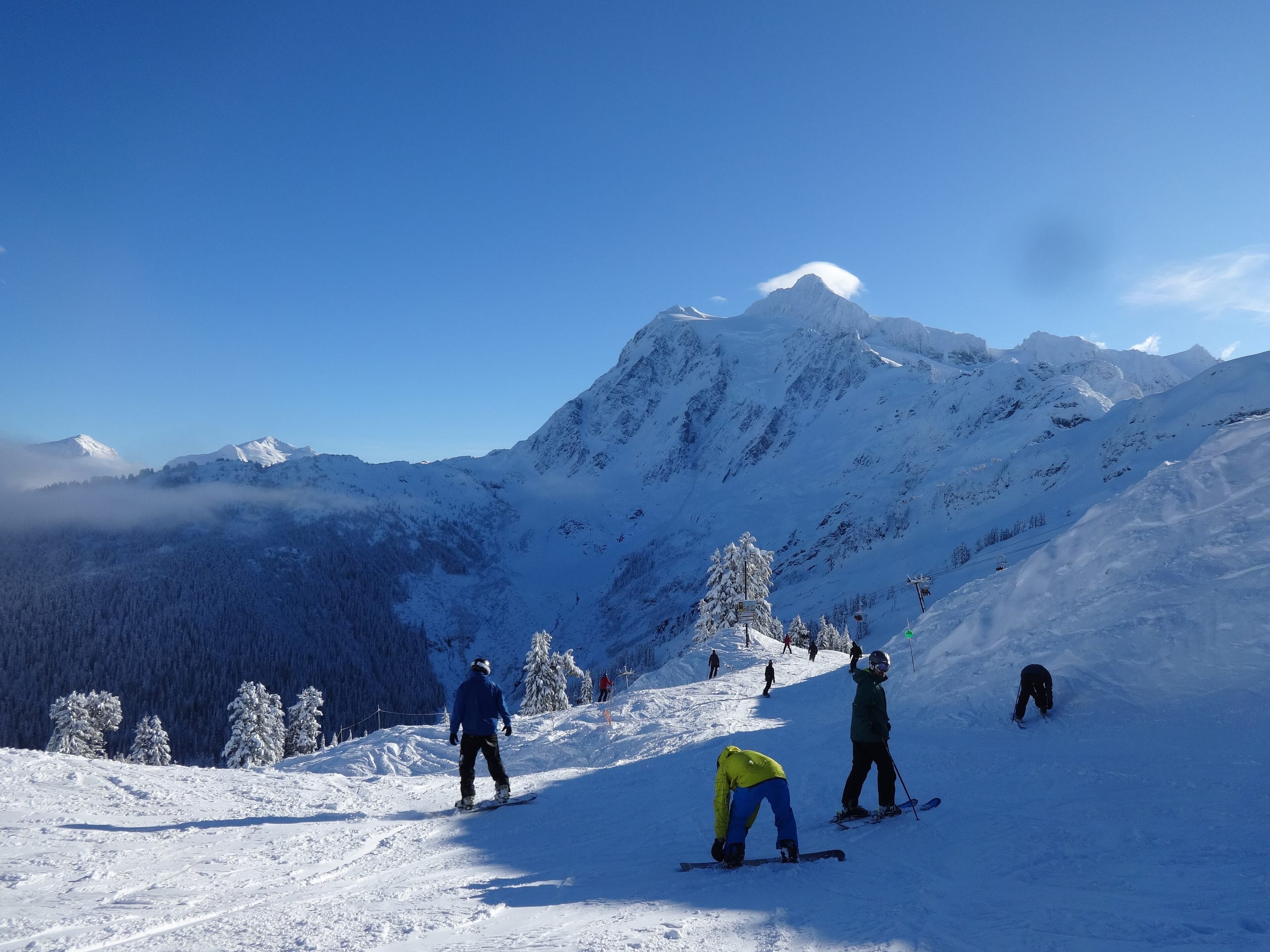 Skiers on a groomed run at Mt. Baker Ski Area with a snow-covered peak and forested valley beyond