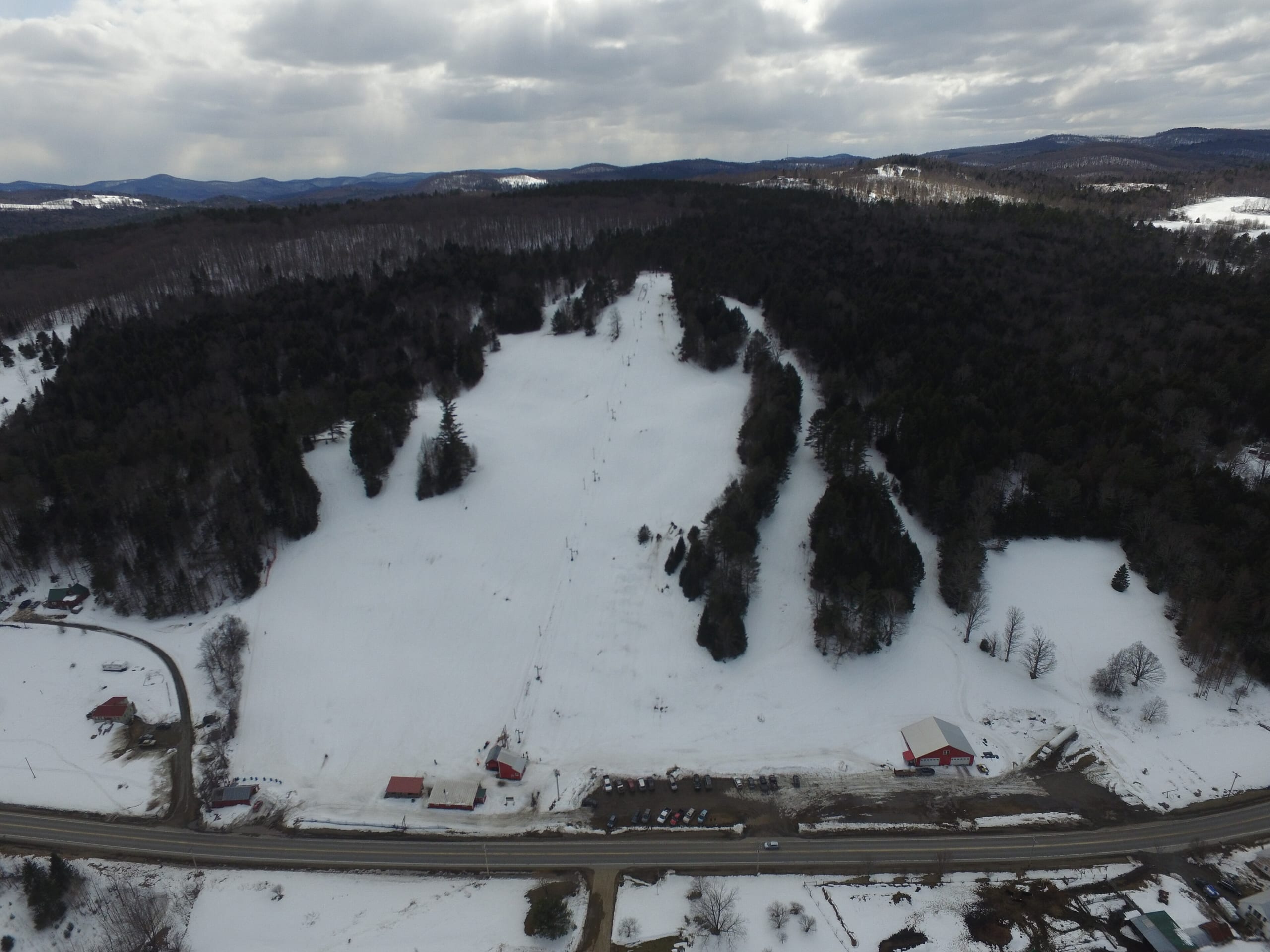 Aerial view of a small snow-covered ski hill with rope tow line, base buildings and a road below