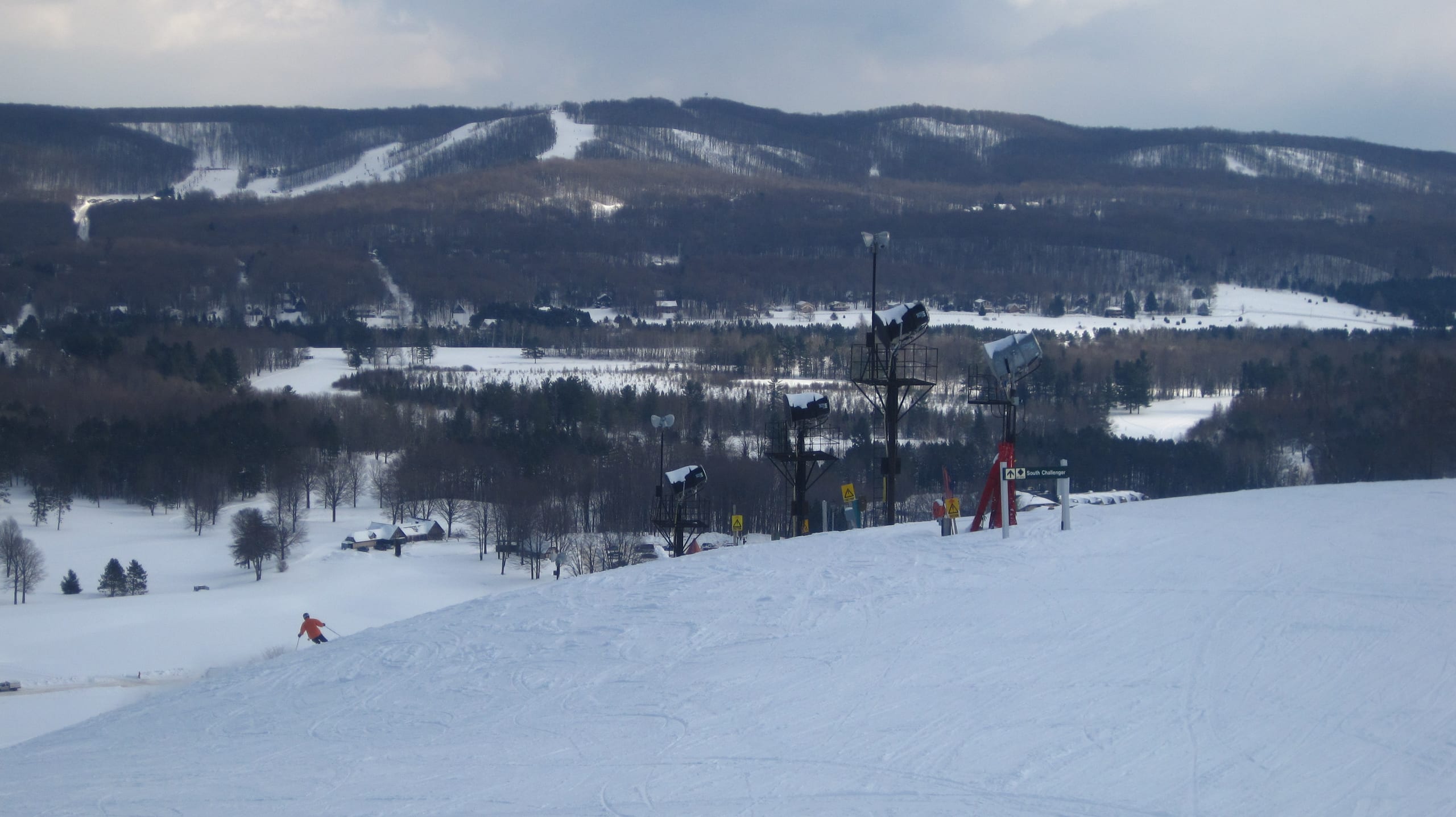 Snowy ski run with snowmaking guns and a lone skier, with wooded hills and trails across the valley