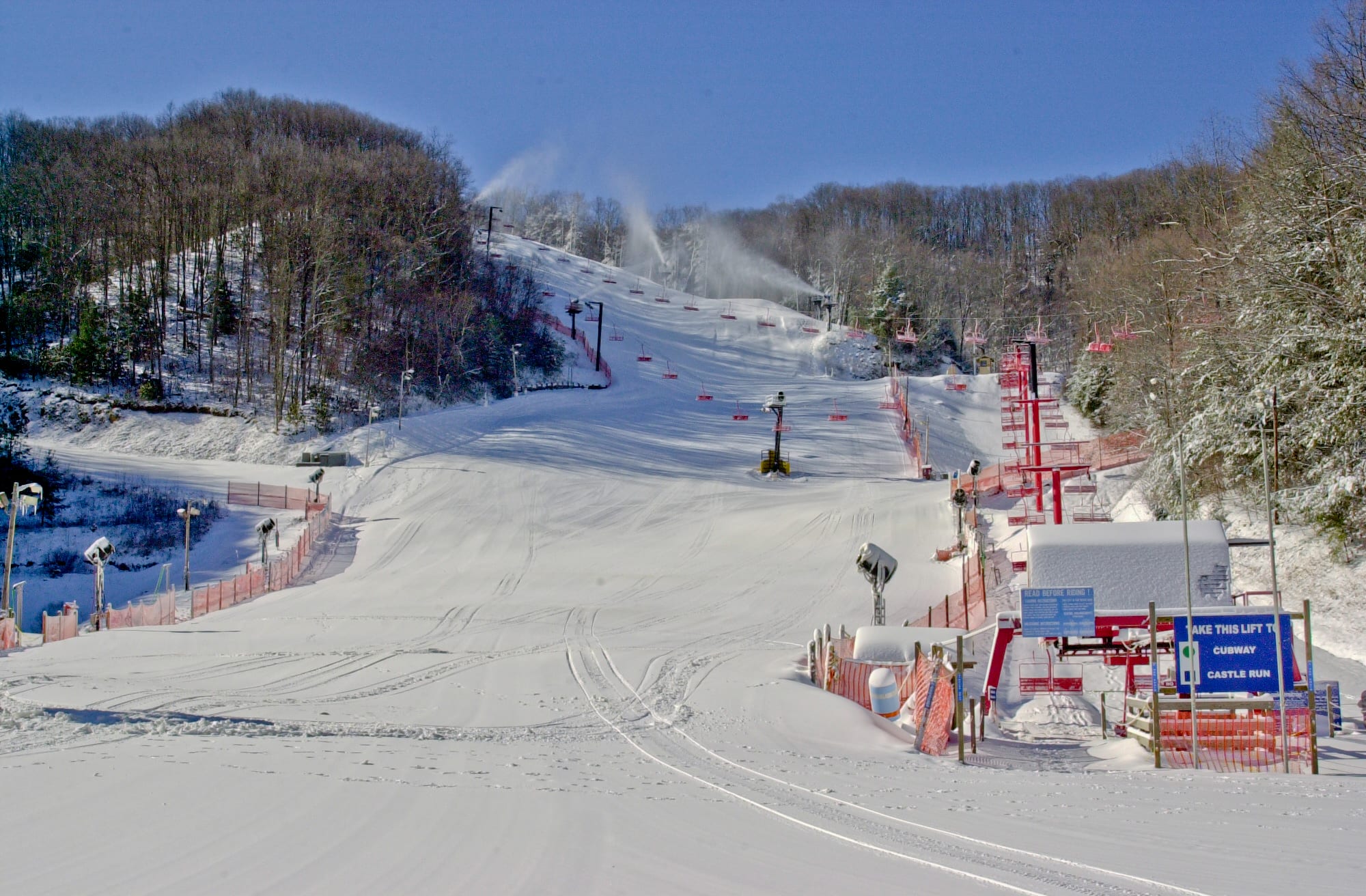 Snow-covered ski slope with snowmaking guns, orange fencing and a chairlift base sign at Ober Gatlinburg