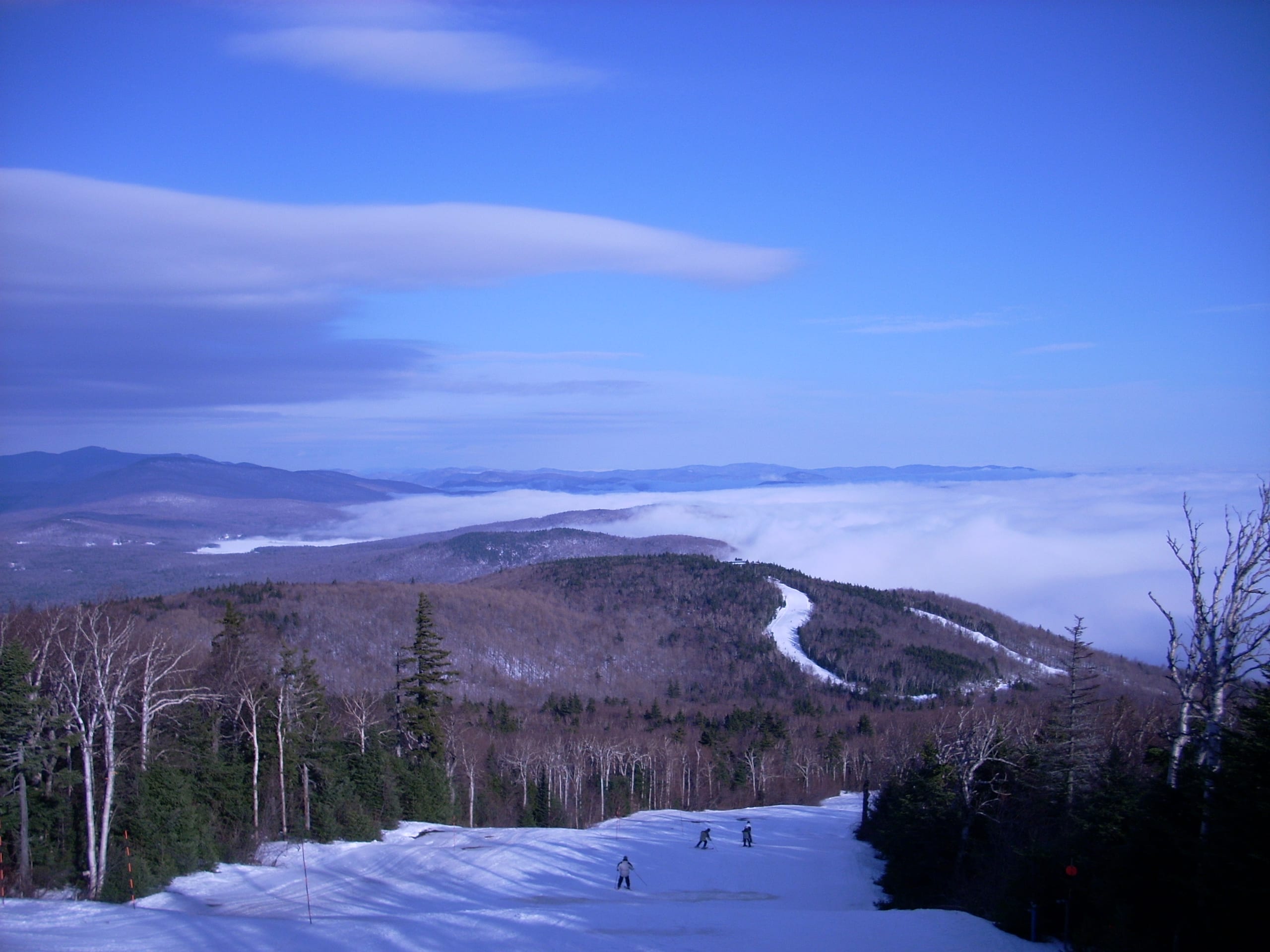 Skiers descend a snowy trail from a wooded summit above a valley filled with clouds in Vermont