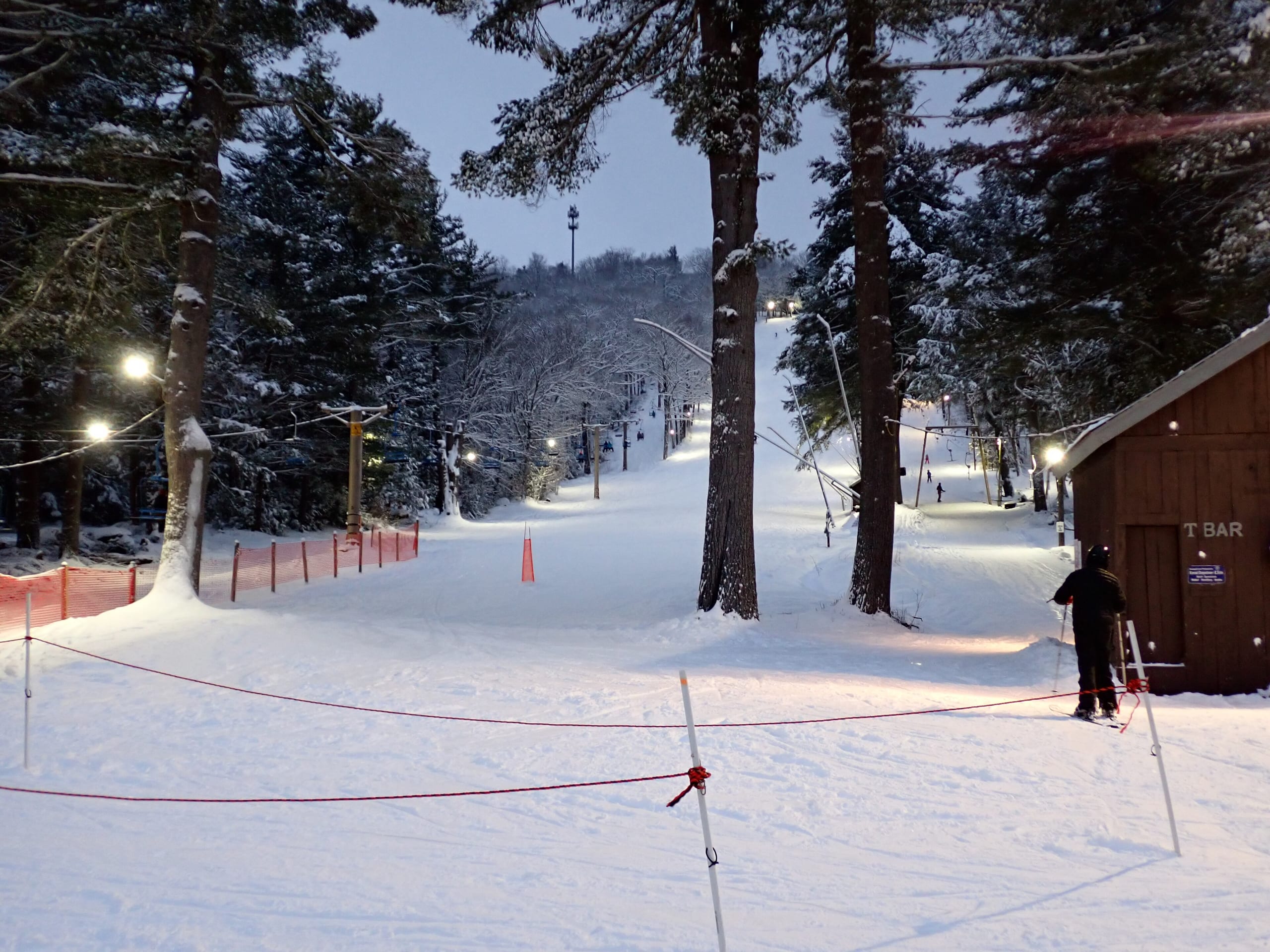 Snow-covered slope lit at dusk with a T-bar shed and skier at the base of a small ski area