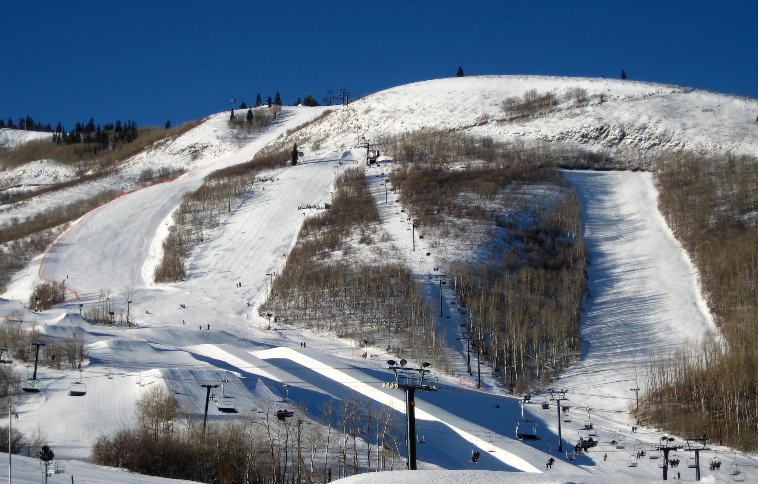 Snow-covered race runs, halfpipe and chairlifts on a broad ski hill under a clear blue sky