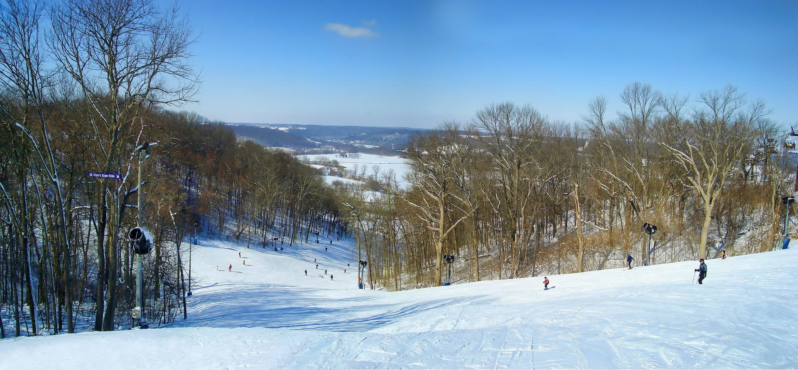 Skiers descend a wide snowy trail lined with bare trees above a river valley at Perfect North Slopes