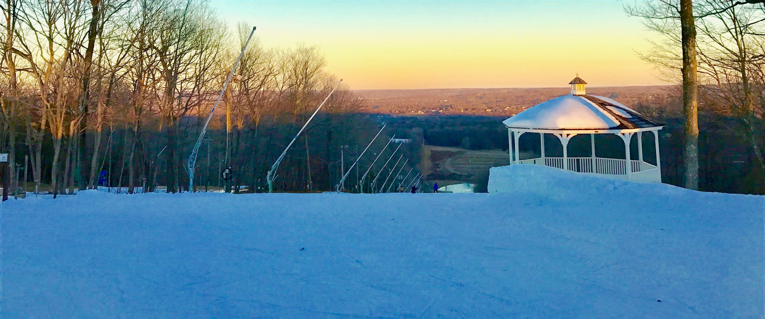 Snowy ski slope at sunset with snowmaking guns along the treeline and a white gazebo on the right.