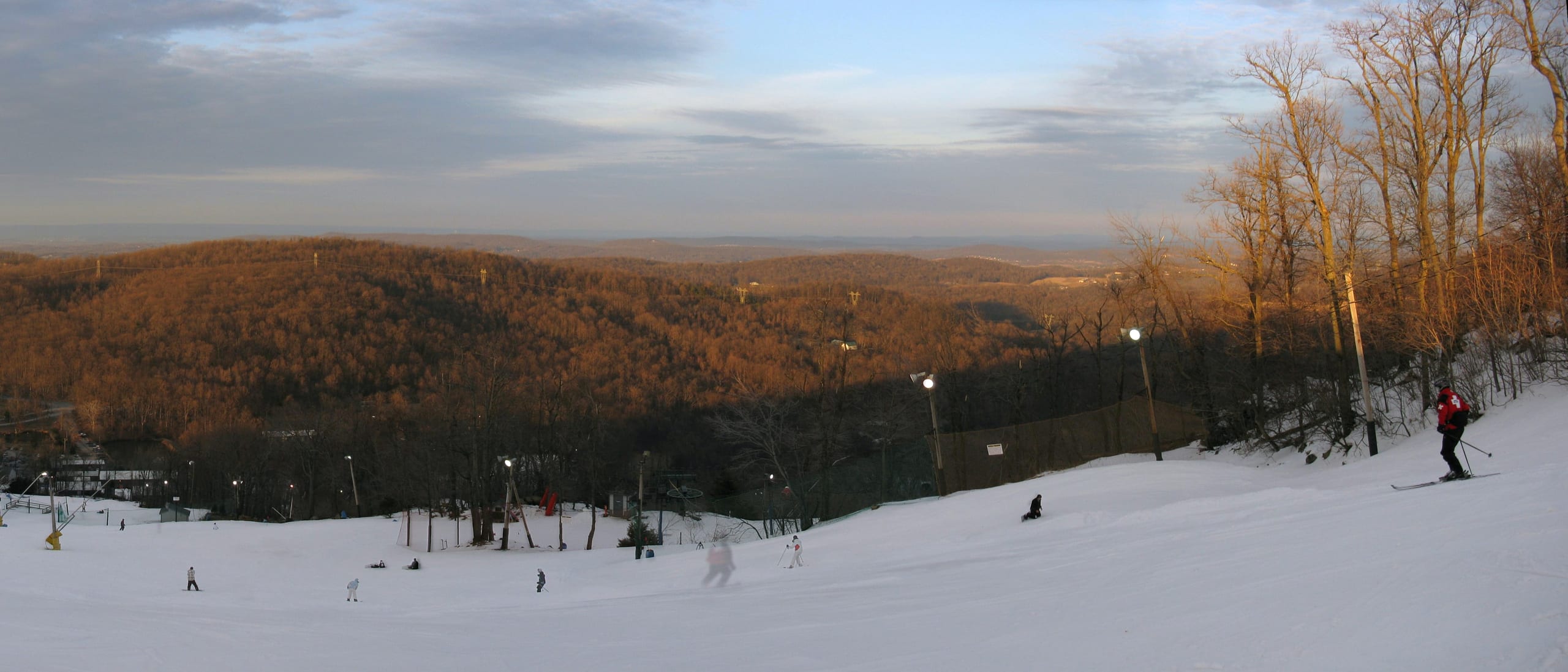 Skiers on a floodlit snow run at Ski Roundtop at dusk, with bare forested ridges beyond.