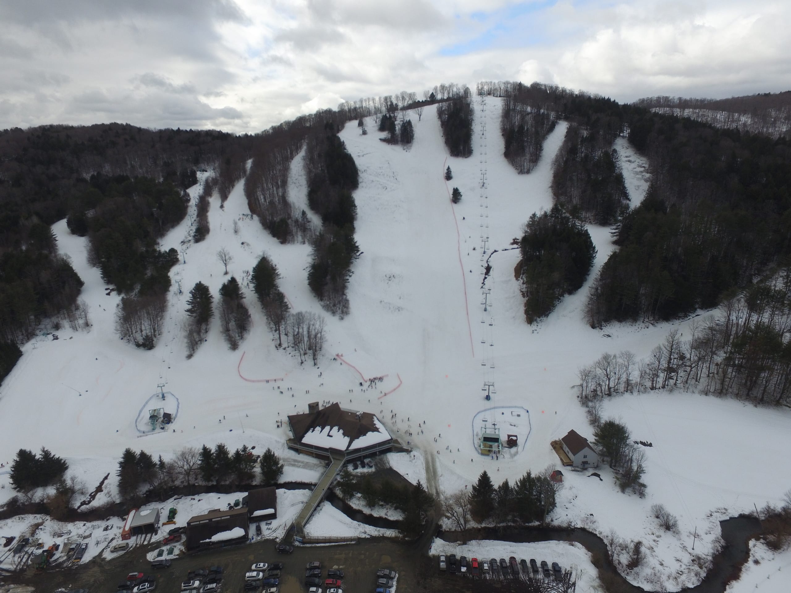 Aerial view of a small ski hill with snowy trails, two lifts, base lodge and parking lot in winter
