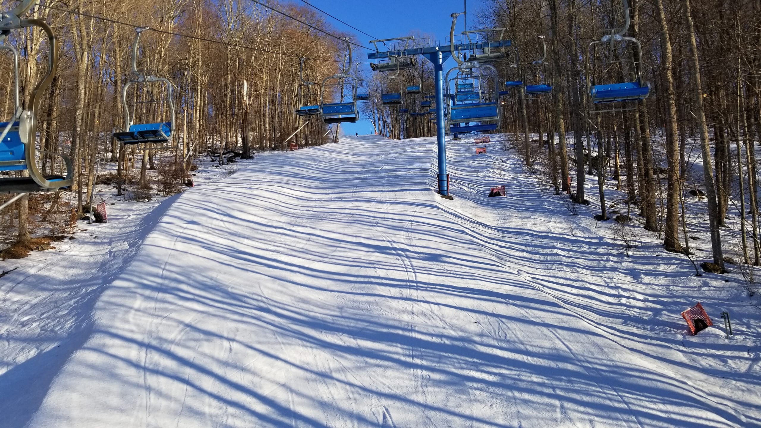 Snow-covered ski trail lined by bare trees with a blue chairlift running overhead